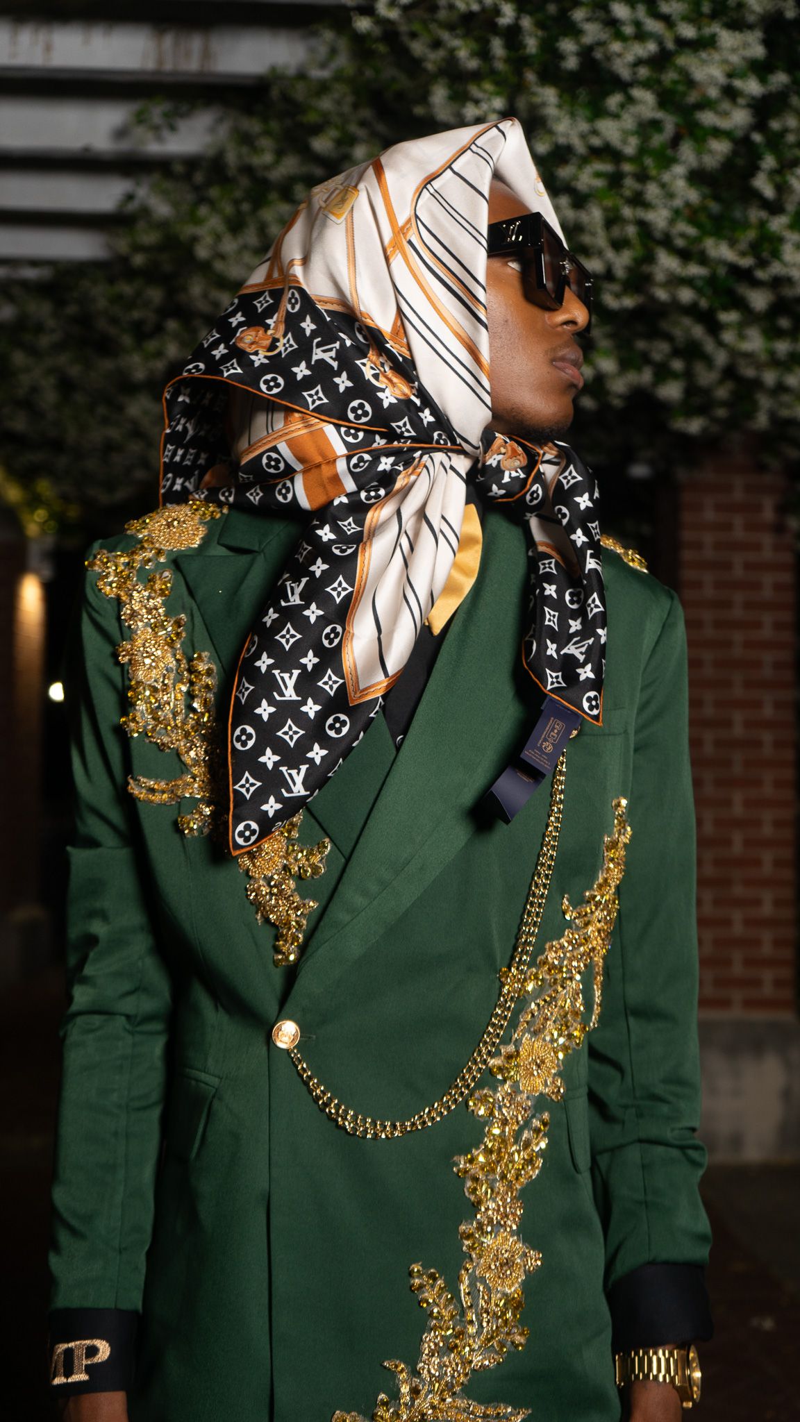 Man in green gold-embroidered blazer with Louis Vuitton scarf headwrap, chain accessory, and sunglasses.