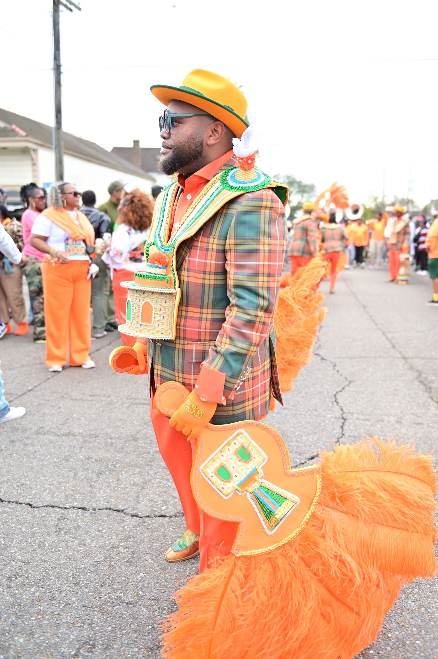 Street performer in plaid jacket and orange pants poses with feathered costume elements.