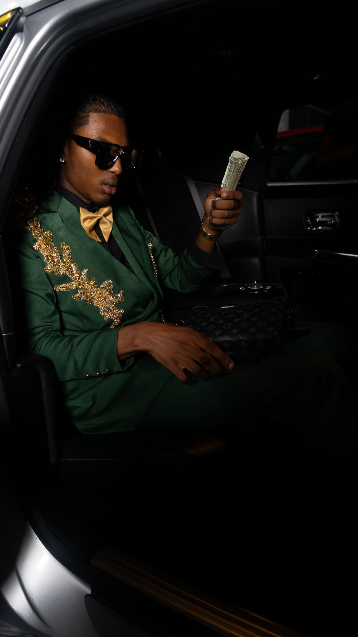 Man in green gold-embroidered suit with bow tie holds cash while seated in luxury car at night.