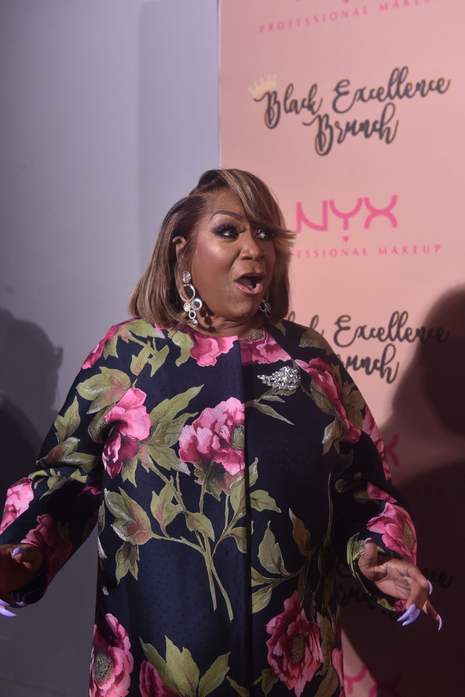 Woman in floral dress poses at NYX Professional Makeup Black Excellence Brunch event backdrop.