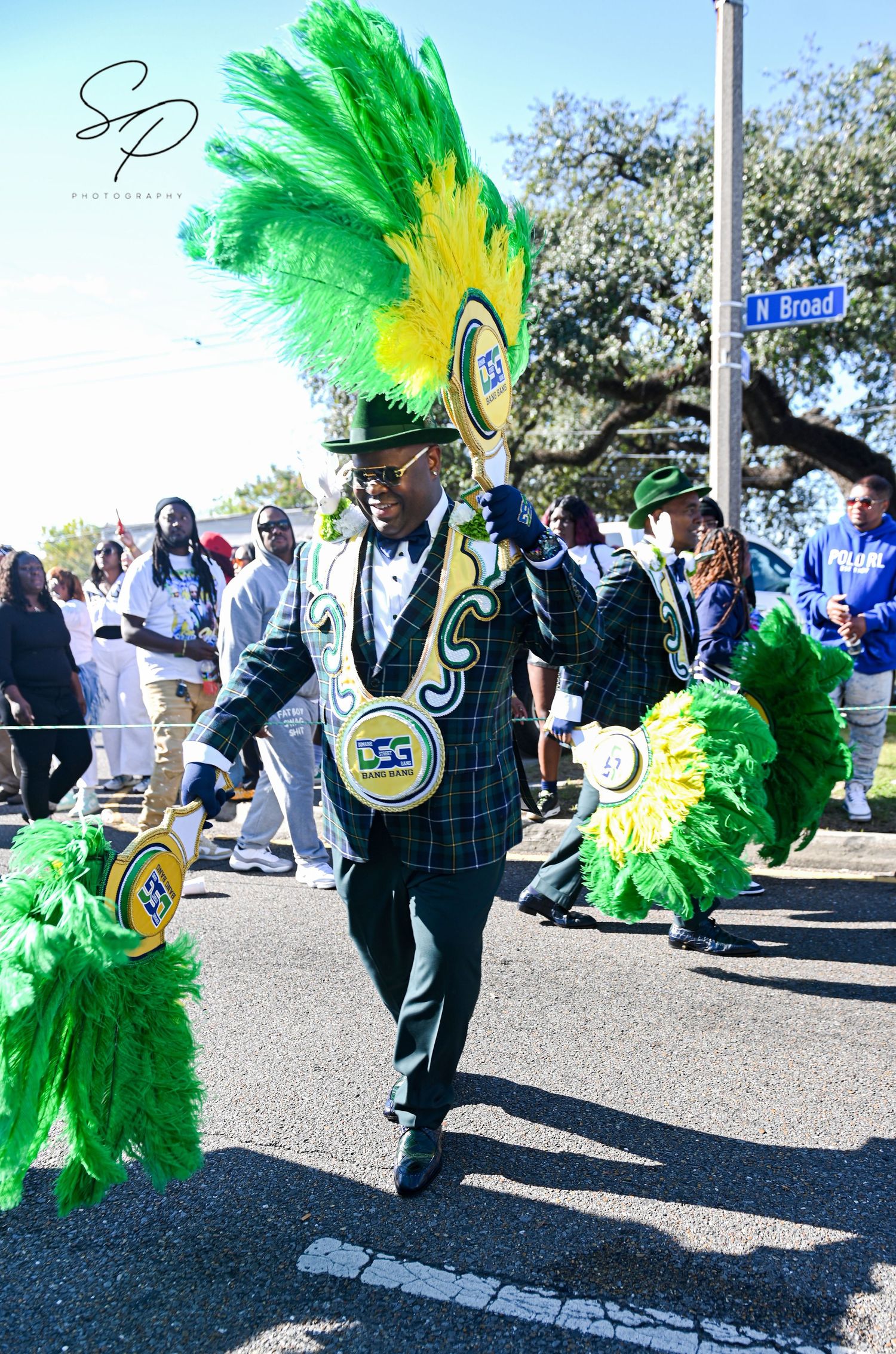 Performers in green feathered costumes and plaid uniforms dancing at a street parade.