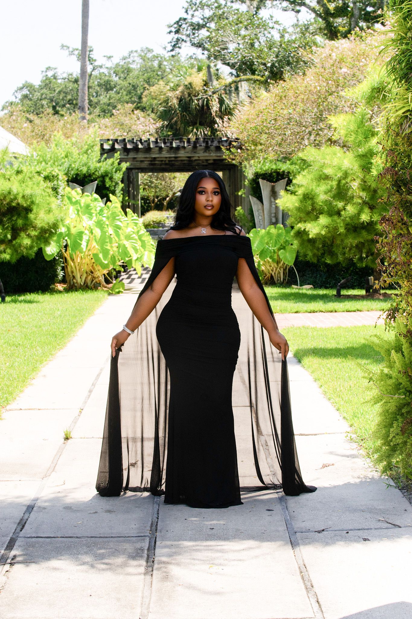 Woman in elegant black off-shoulder cape gown poses on garden pathway surrounded by lush green trees.