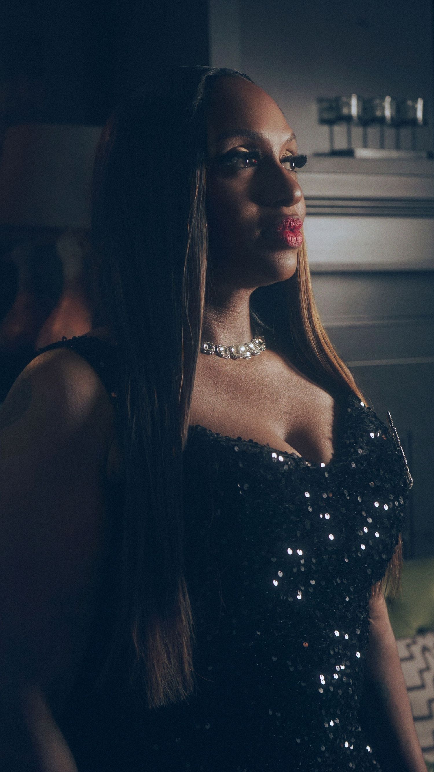 A glamorous woman in a black sequin dress with a diamond choker necklace and long straight hair poses in dim lighting.