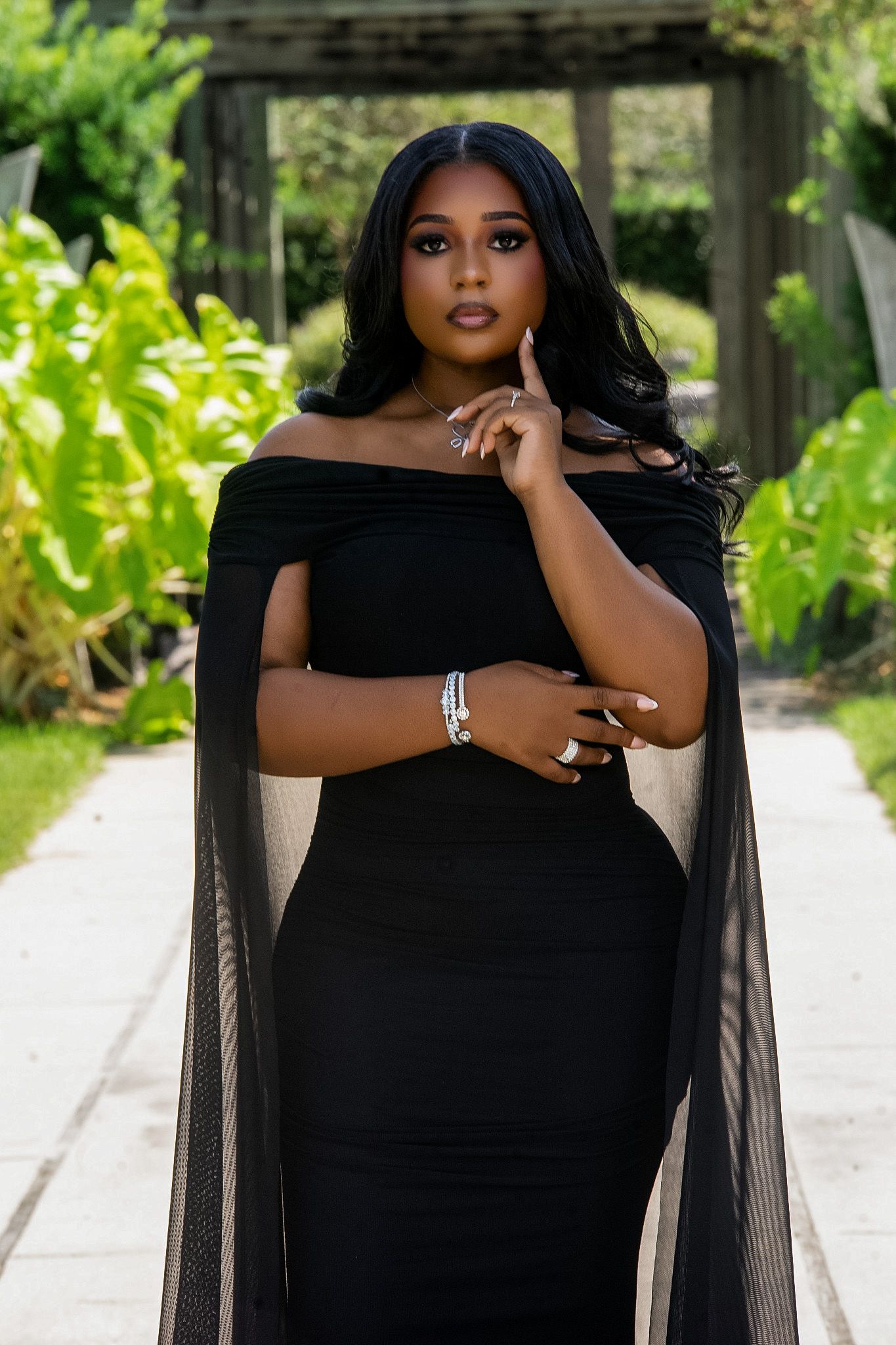 Woman in black off-shoulder cape gown poses gracefully in a lush garden, wearing silver jewelry.