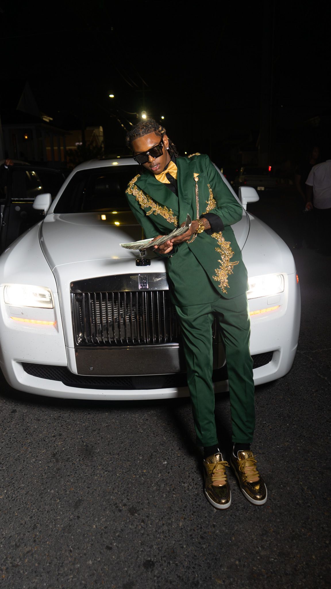 Man in green and gold embroidered tracksuit leaning on white Rolls-Royce holding cash at night.