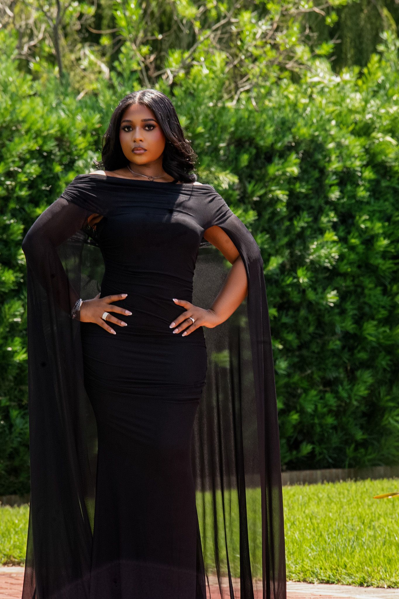 Woman in elegant black off-shoulder gown with sheer cape sleeves poses outdoors against lush green hedges.