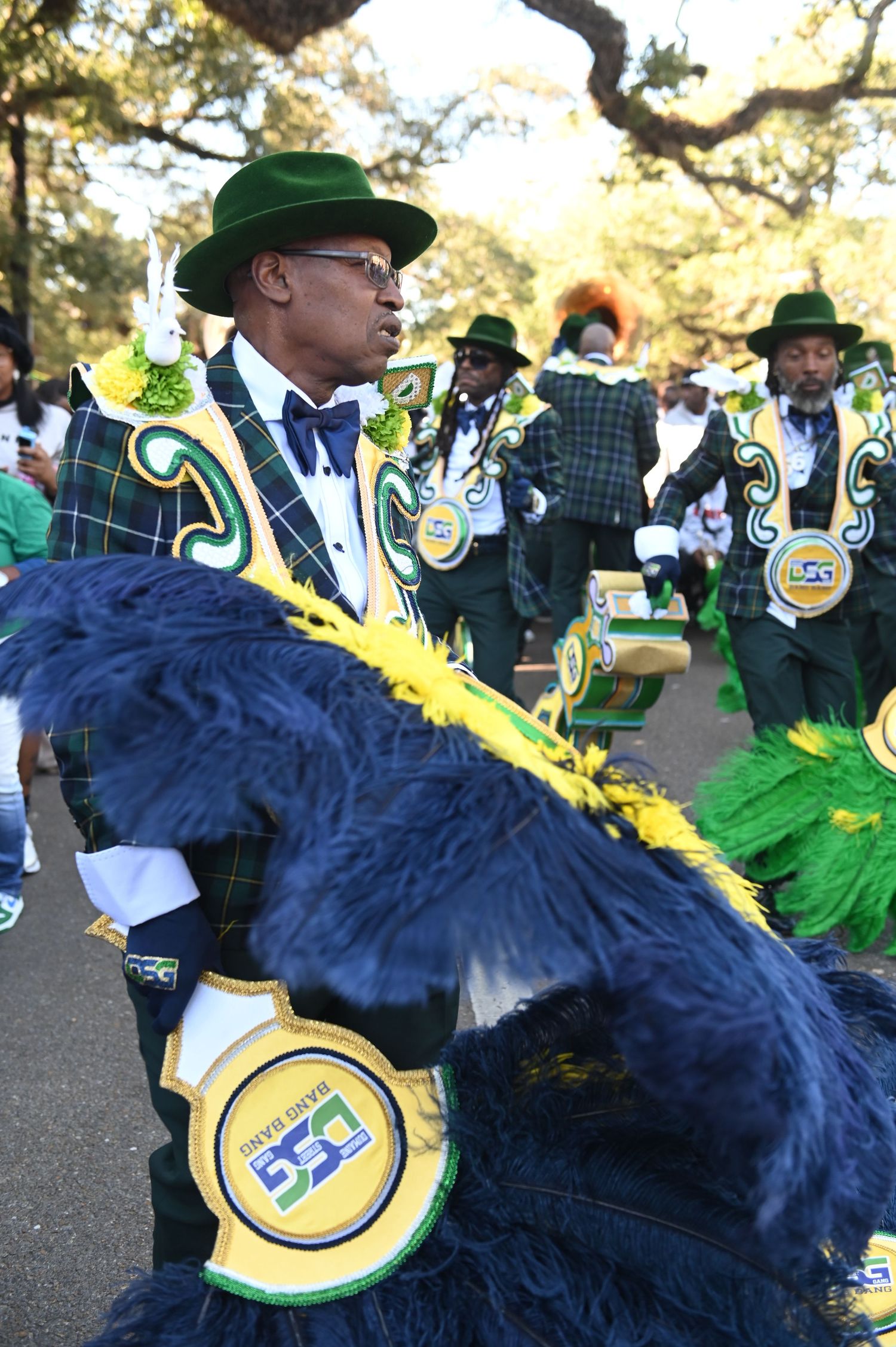 Marching band performers in green and blue plaid uniforms with feathered accessories perform during a parade celebration.