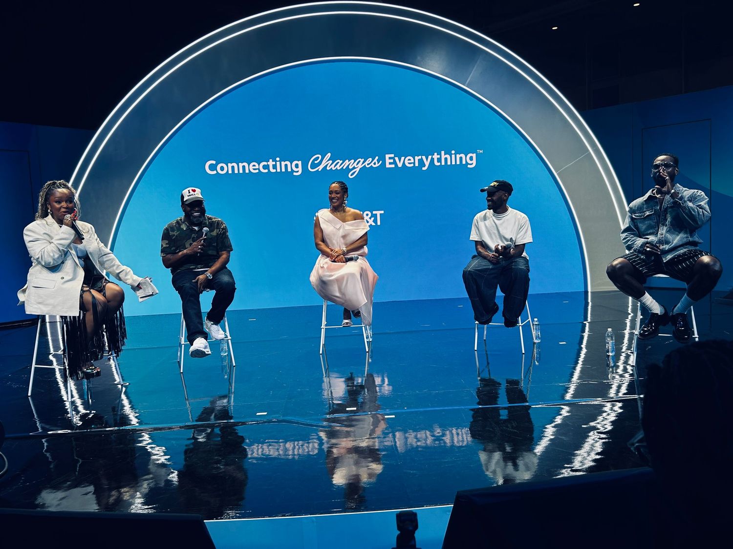 Panel discussion on stage with circular blue backdrop featuring 'Connecting Through Everything' text.