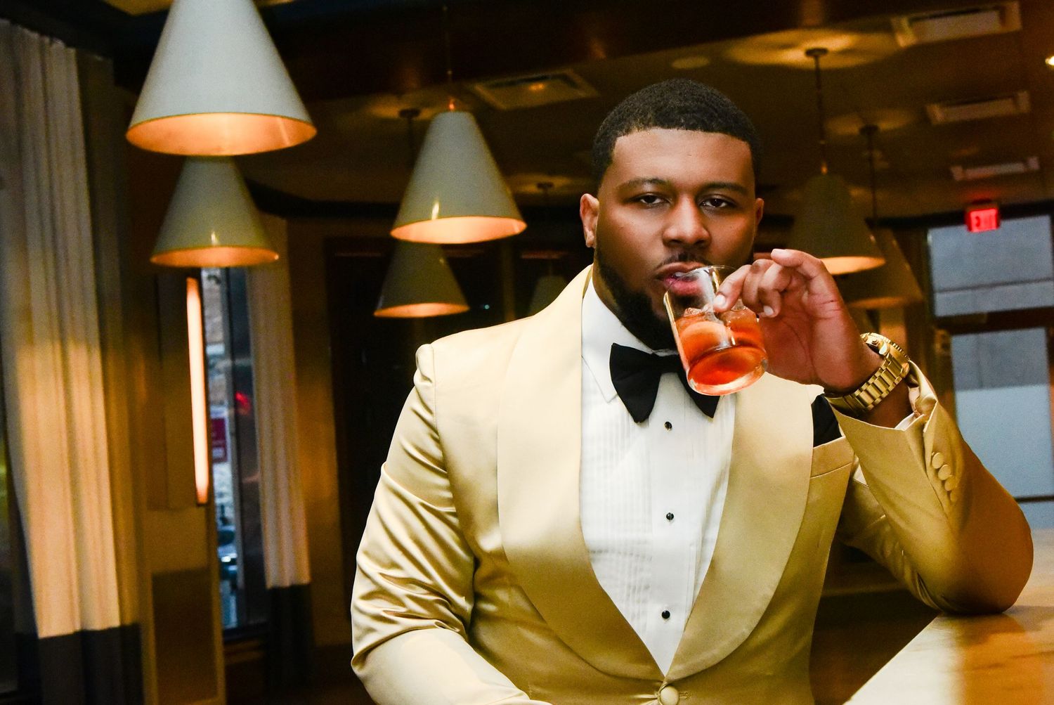 Man in gold tuxedo with bow tie sipping cocktail at upscale bar, pendant lights overhead.
