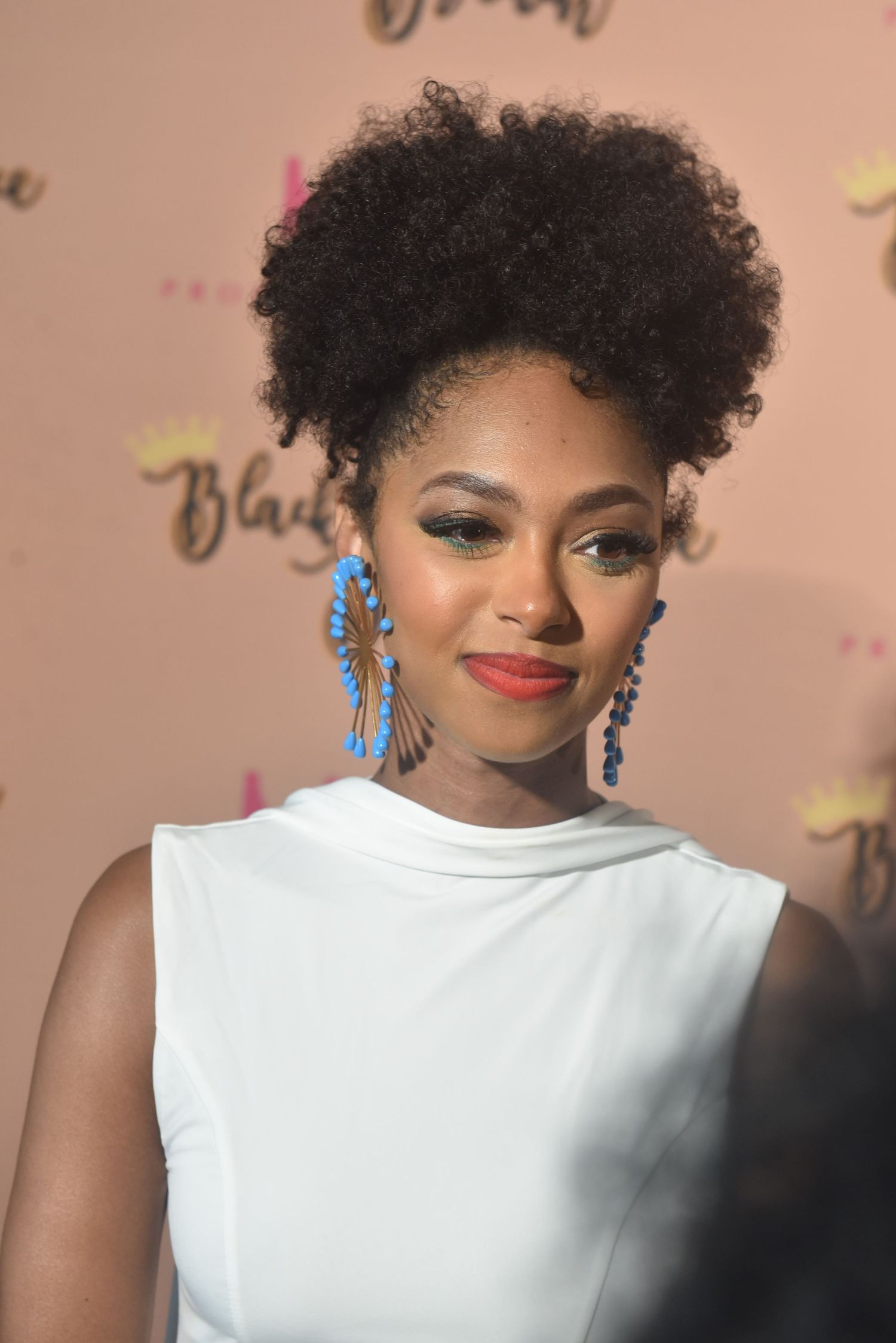 Young woman with natural afro updo wearing blue statement earrings and white sleeveless top at an event.