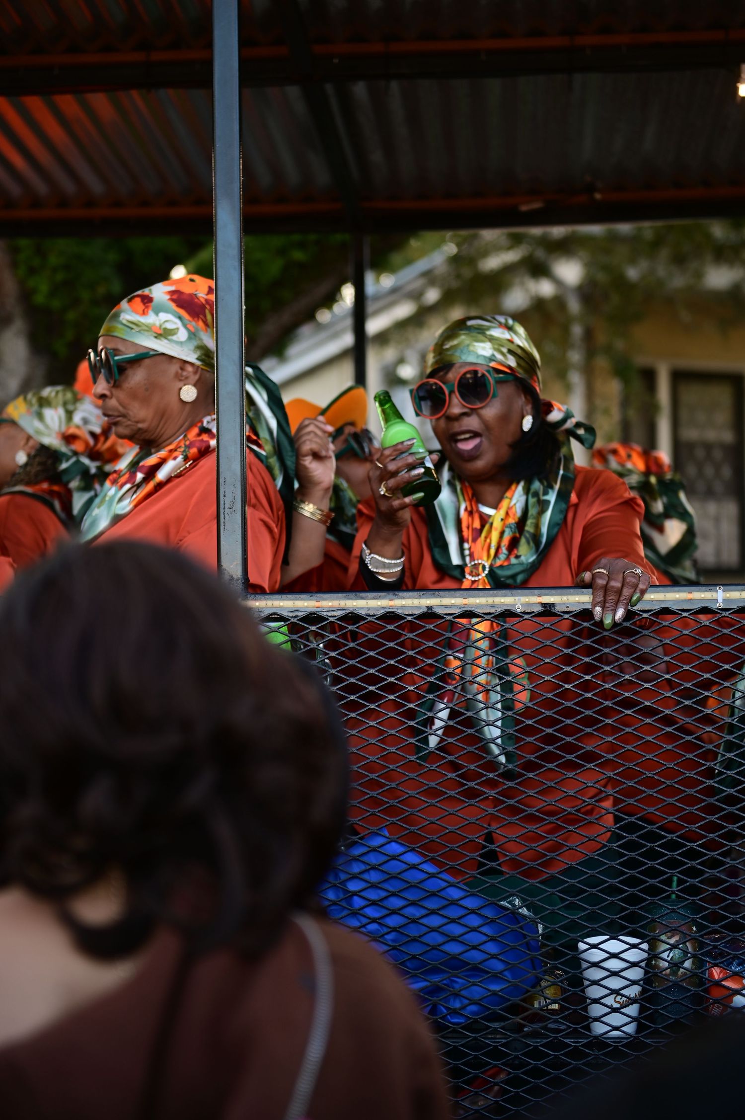 Festival participants in bright orange attire and sunglasses celebrate from behind a barrier during a parade event.