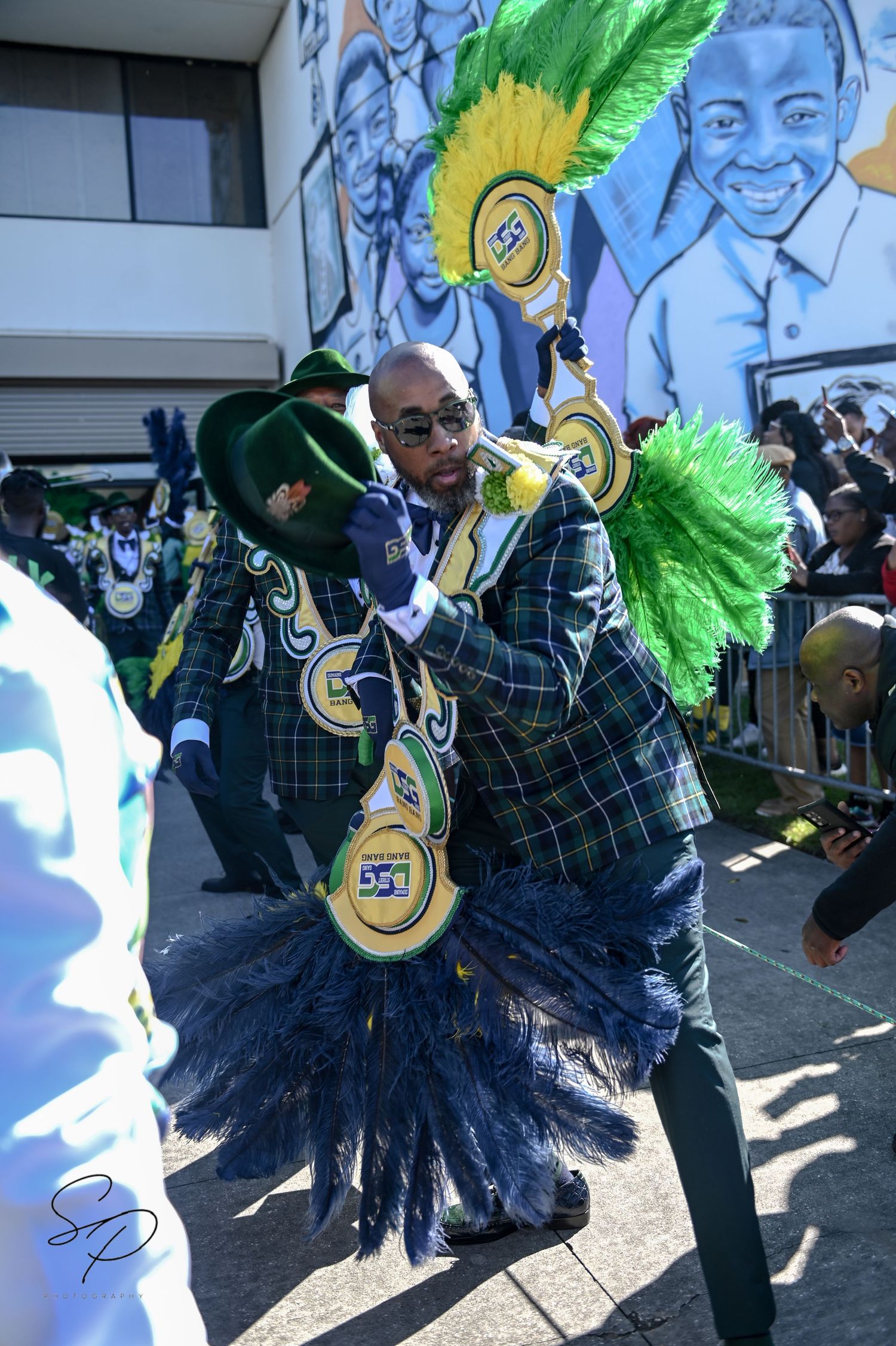 Performer in plaid suit with green and gold feathered accessories during street celebration.