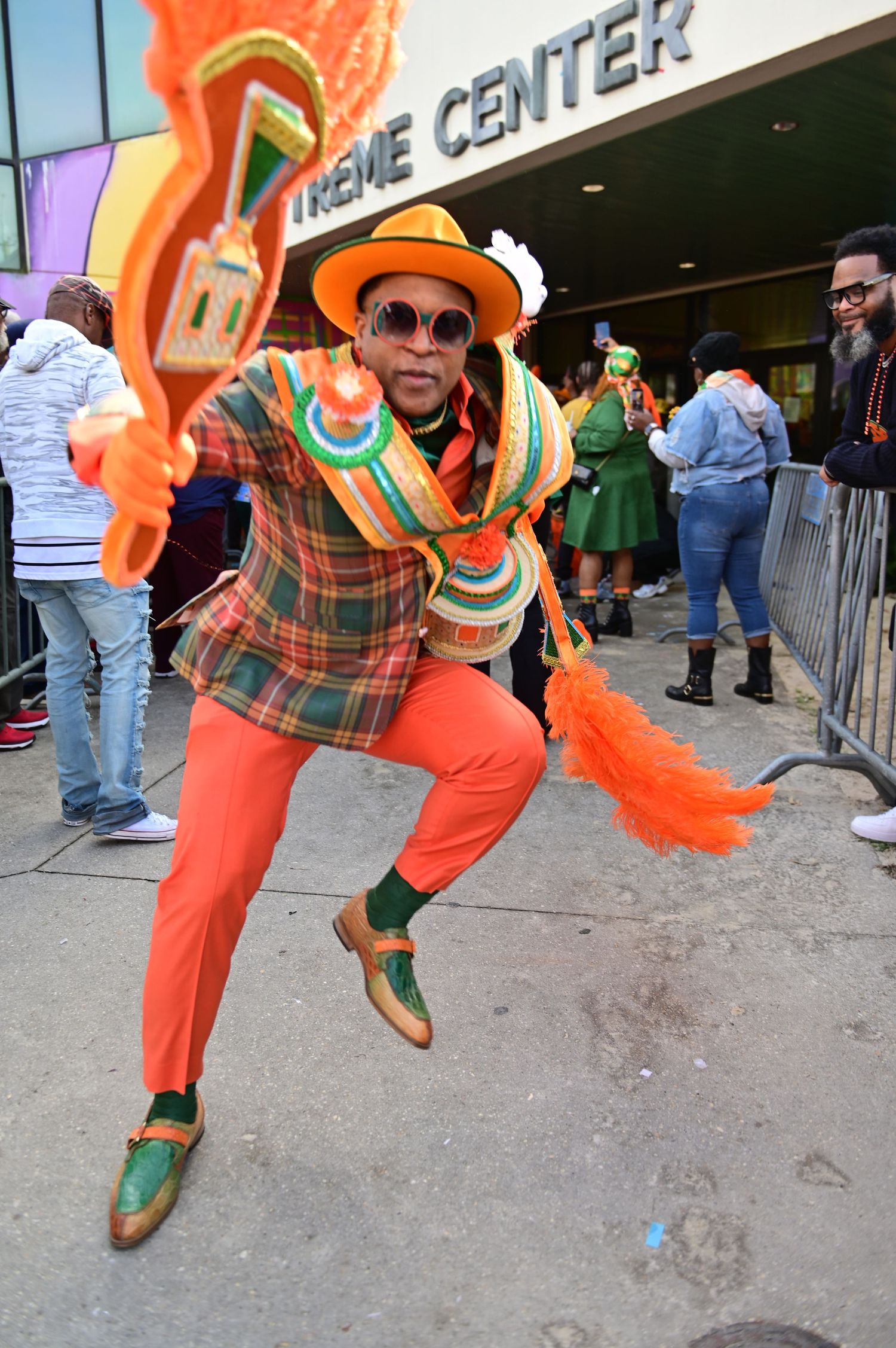 Performer in vibrant plaid suit and orange pants strikes dynamic pose outside venue center.