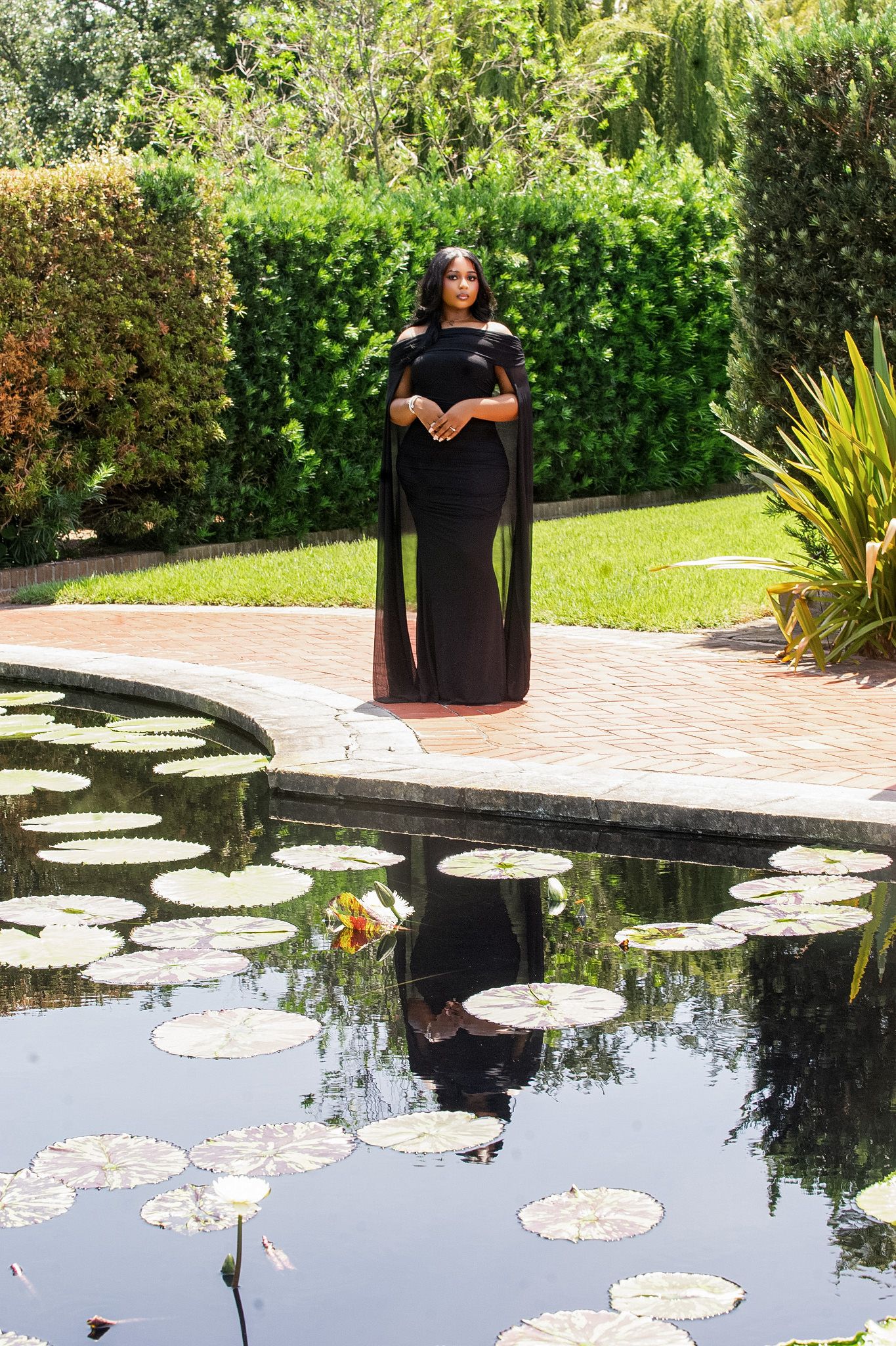 Pregnant woman in elegant black cape gown standing by a lily pad pond in a lush garden setting.