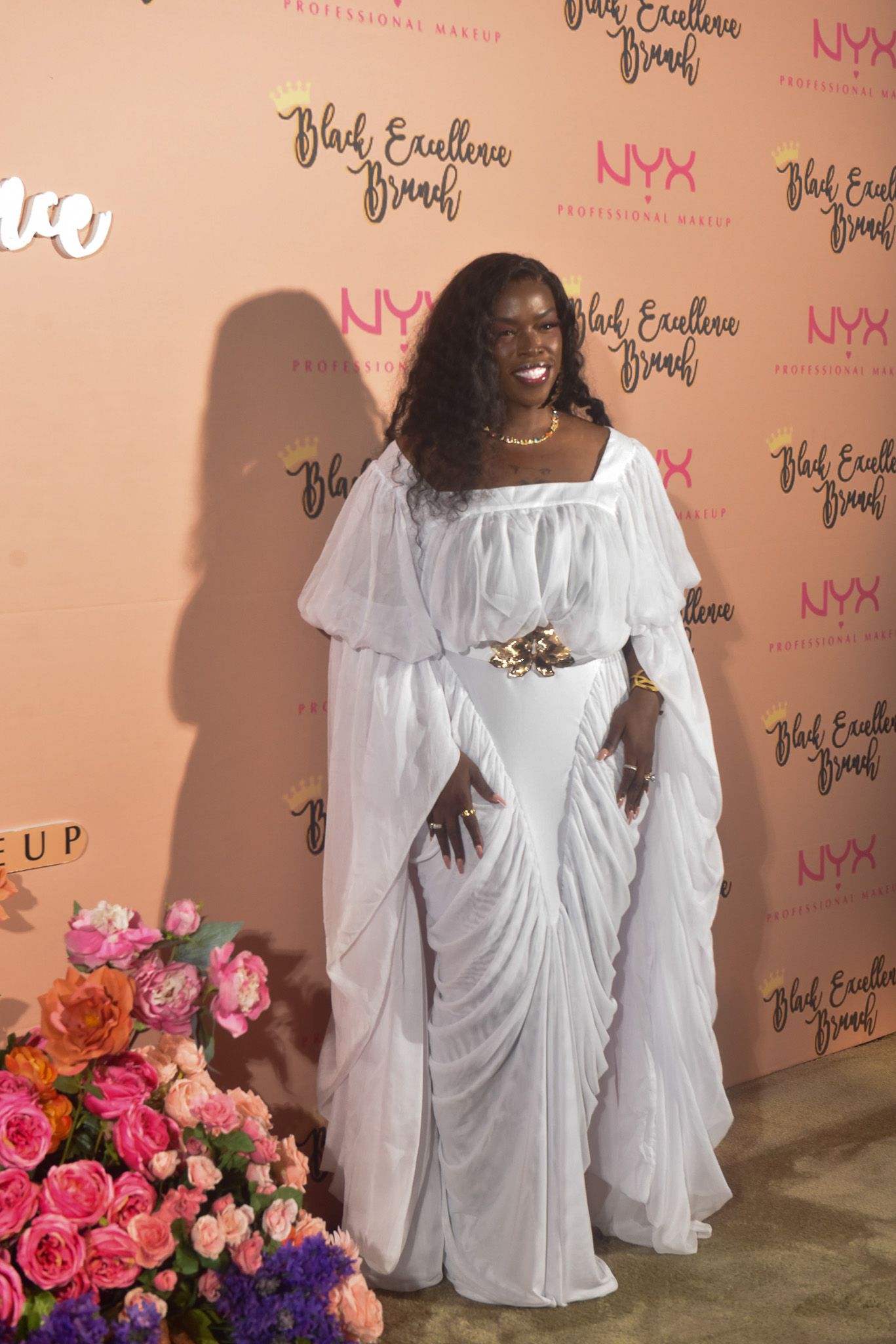 Woman in white flowing gown poses at NYX Professional Makeup Black Excellence Brunch event with floral decor.