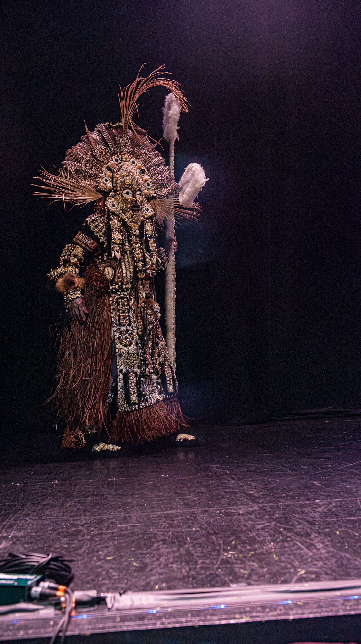 Elaborate tribal costume with feathered headdress and staff on dark stage, showcasing intricate ceremonial attire.