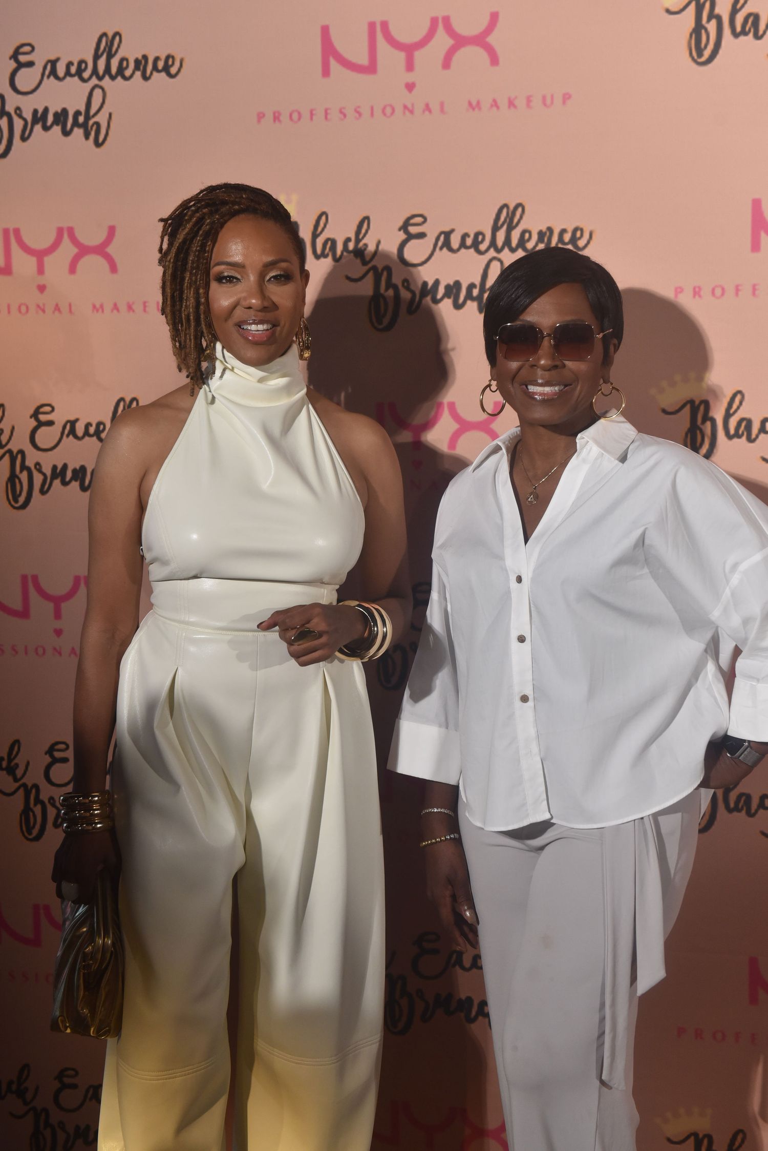 Two women pose together at NYX Professional Makeup Black Excellence Brunch event, both wearing elegant white outfits.