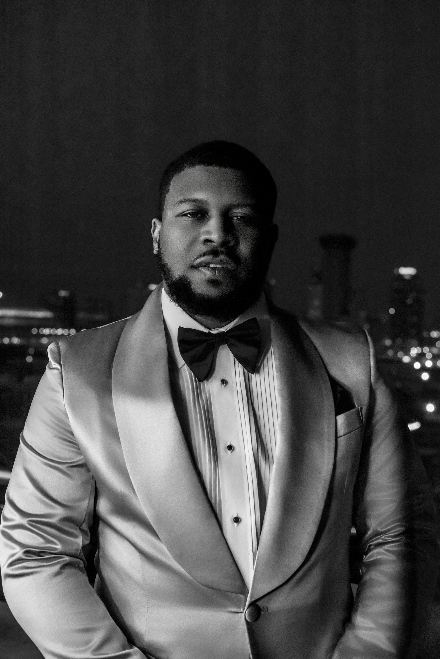 A man in a white tuxedo with bow tie poses confidently against a city night skyline backdrop.