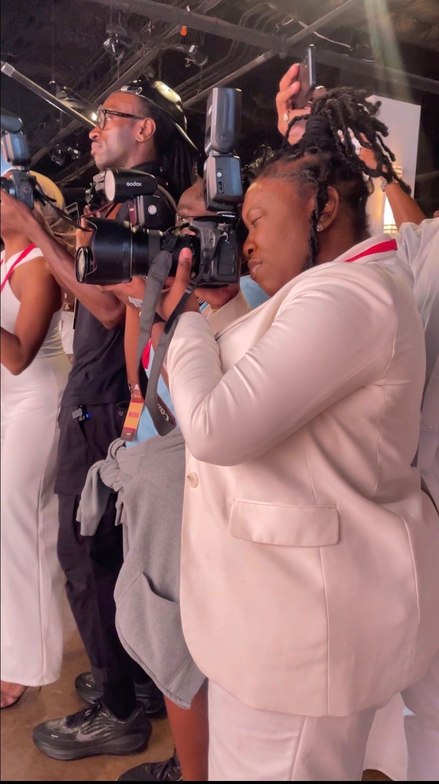Two photographers in white attire capture shots at an indoor event with studio lighting equipment visible.