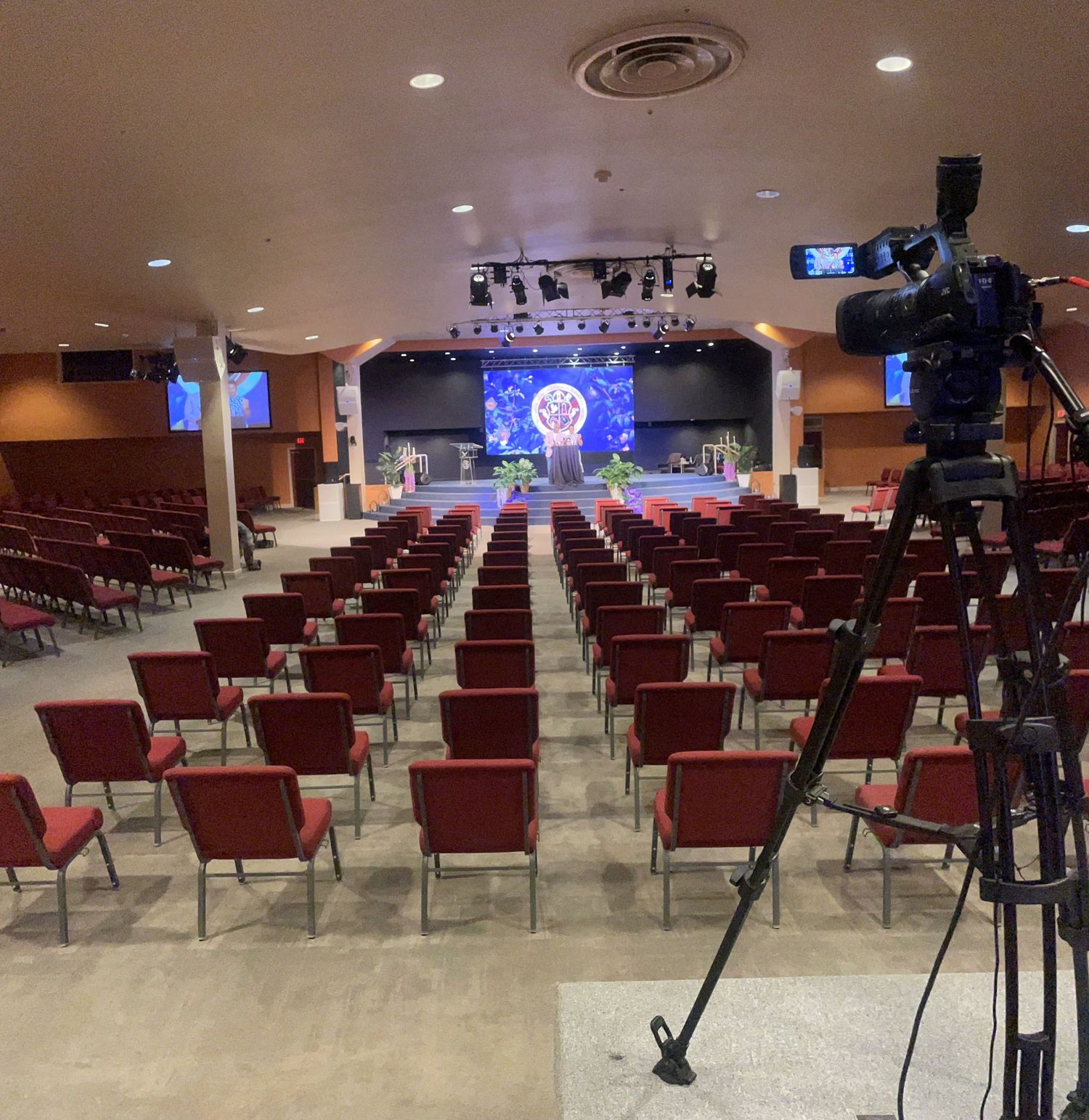Large event hall with red chairs, stage with colorful display, and a professional video camera on tripod in foreground.