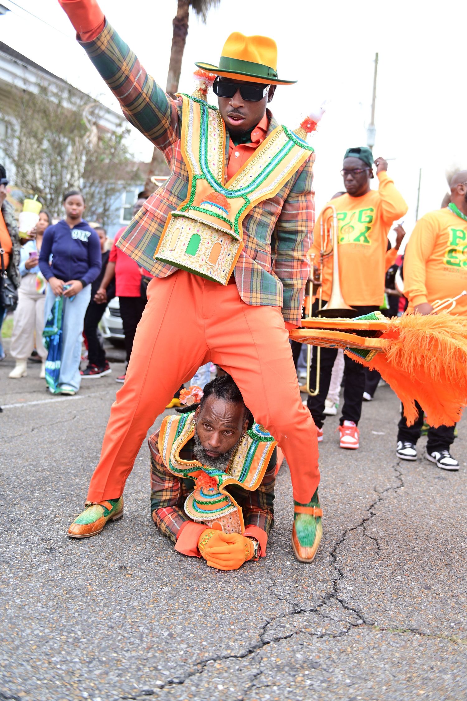 Two men in vibrant orange and green plaid suits perform a stunt at a colorful street parade celebration.