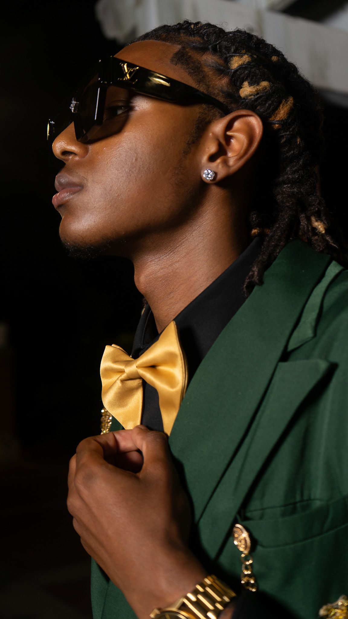 Stylish man in green blazer with gold bow tie, sunglasses, dreadlocks, and gold watch posing confidently.