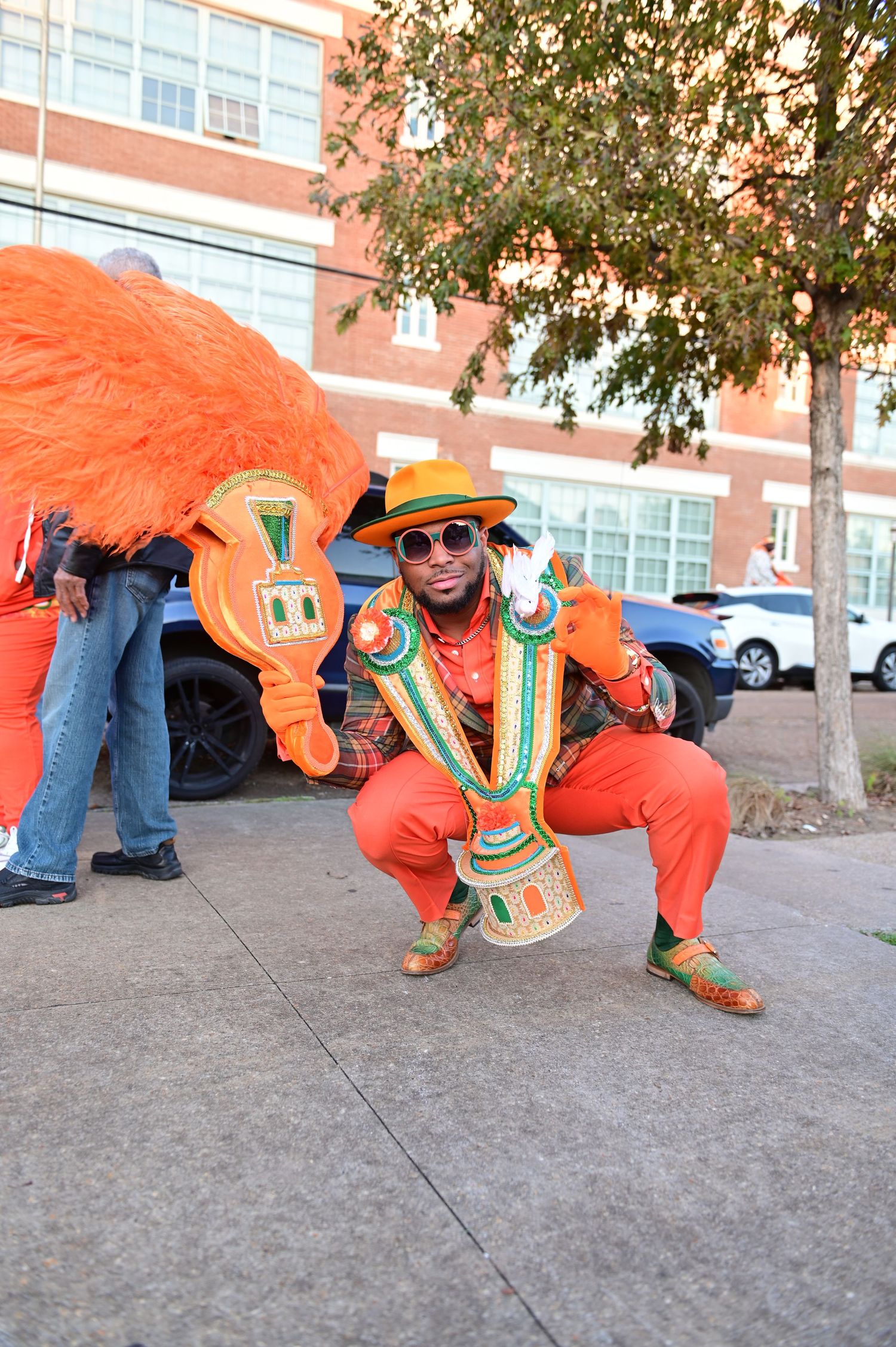 Performer in vibrant orange costume and sunglasses poses with decorative props during a street celebration.