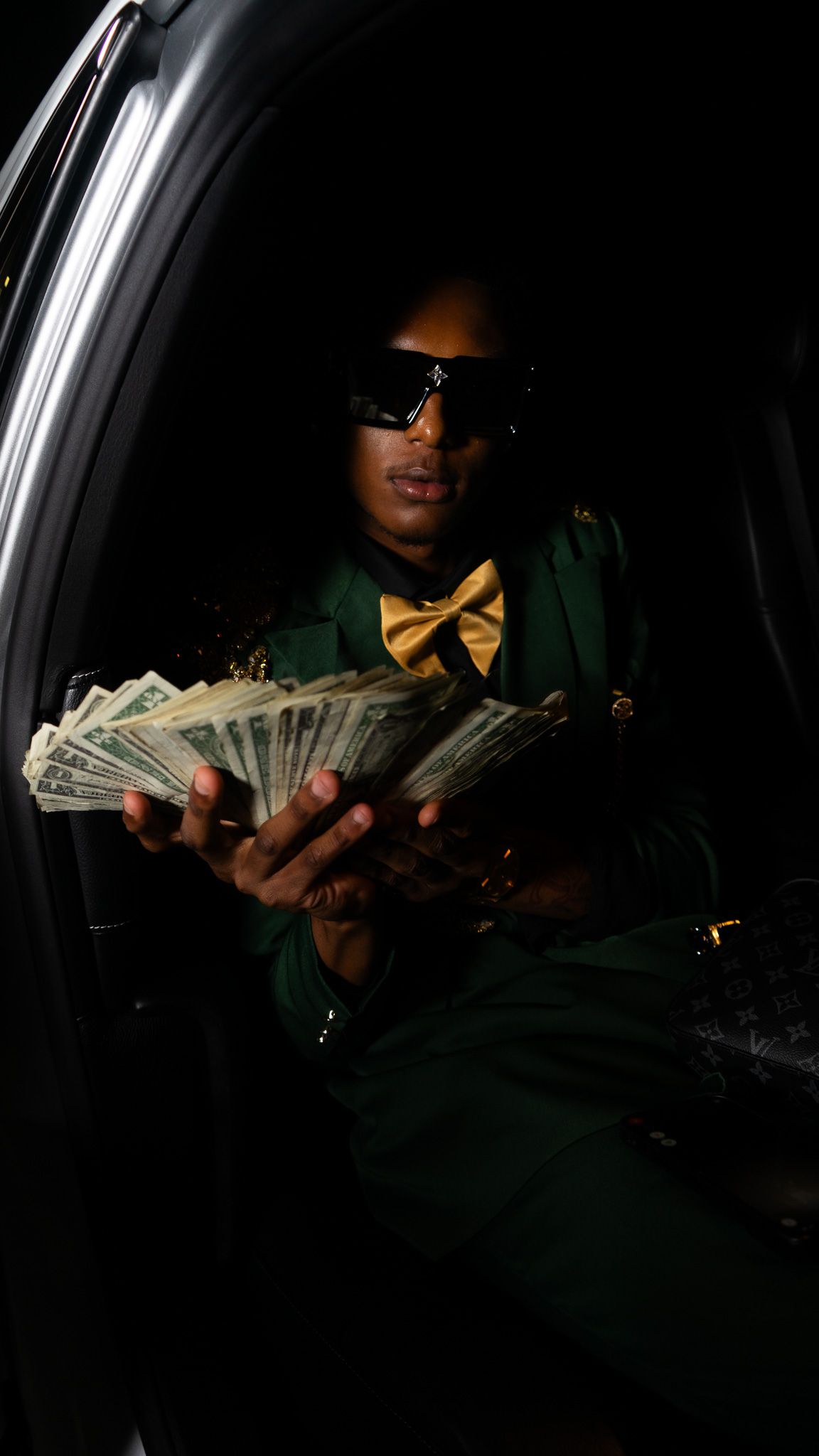 Man in green suit and yellow bow tie holding fanned cash while sitting in a dark car interior.
