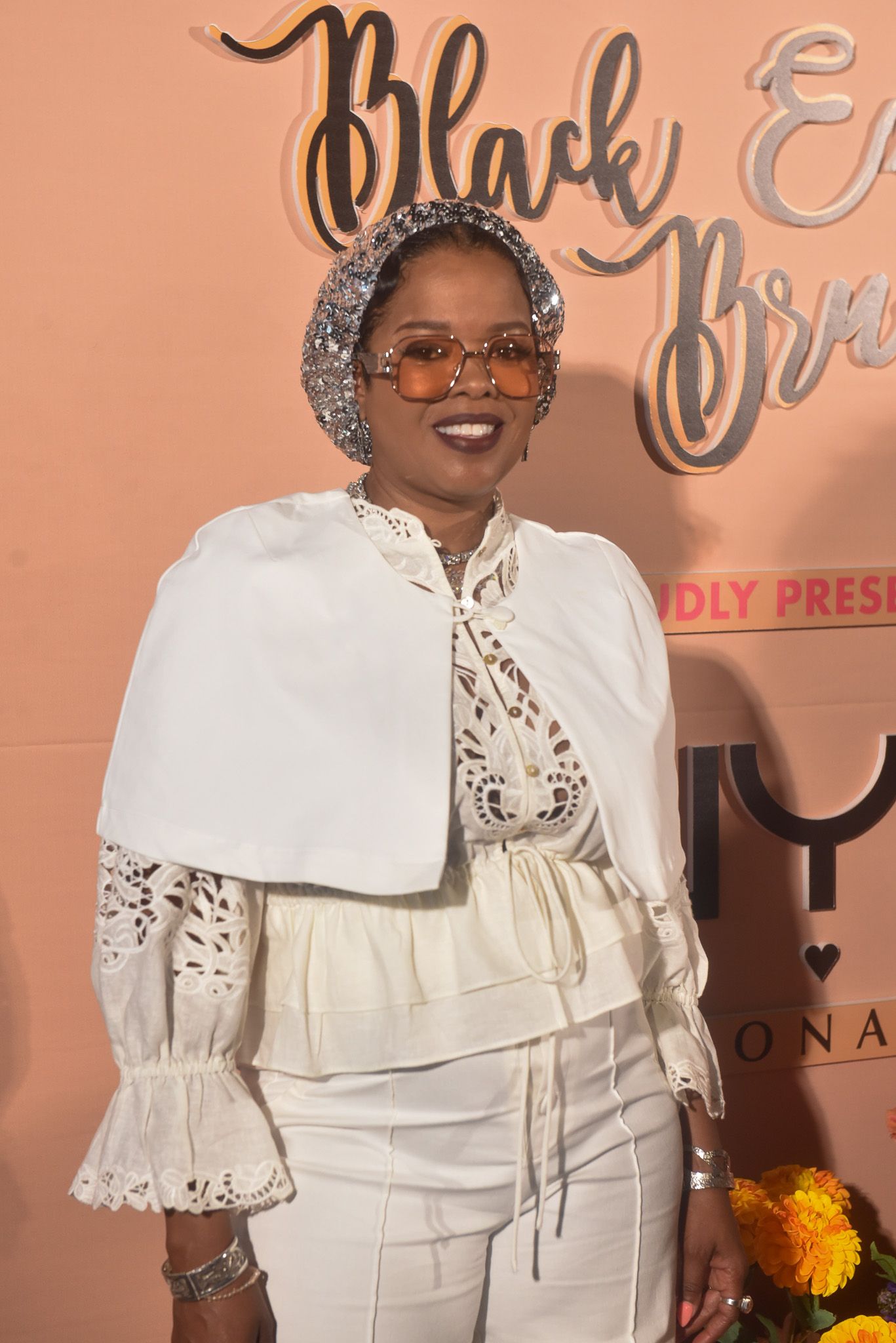 Woman in white lace outfit and silver headscarf poses at Black Excellence Brunch event backdrop.