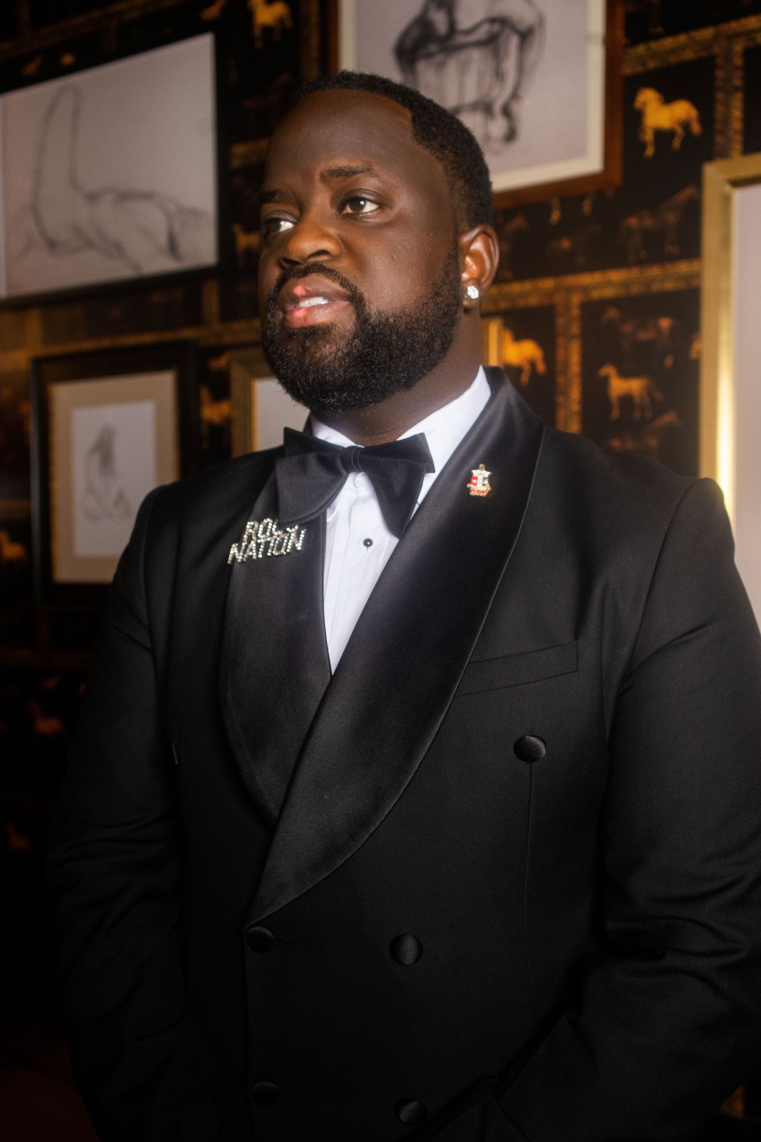 Man in black tuxedo with bow tie and lapel pins poses at formal event with framed artwork in background.