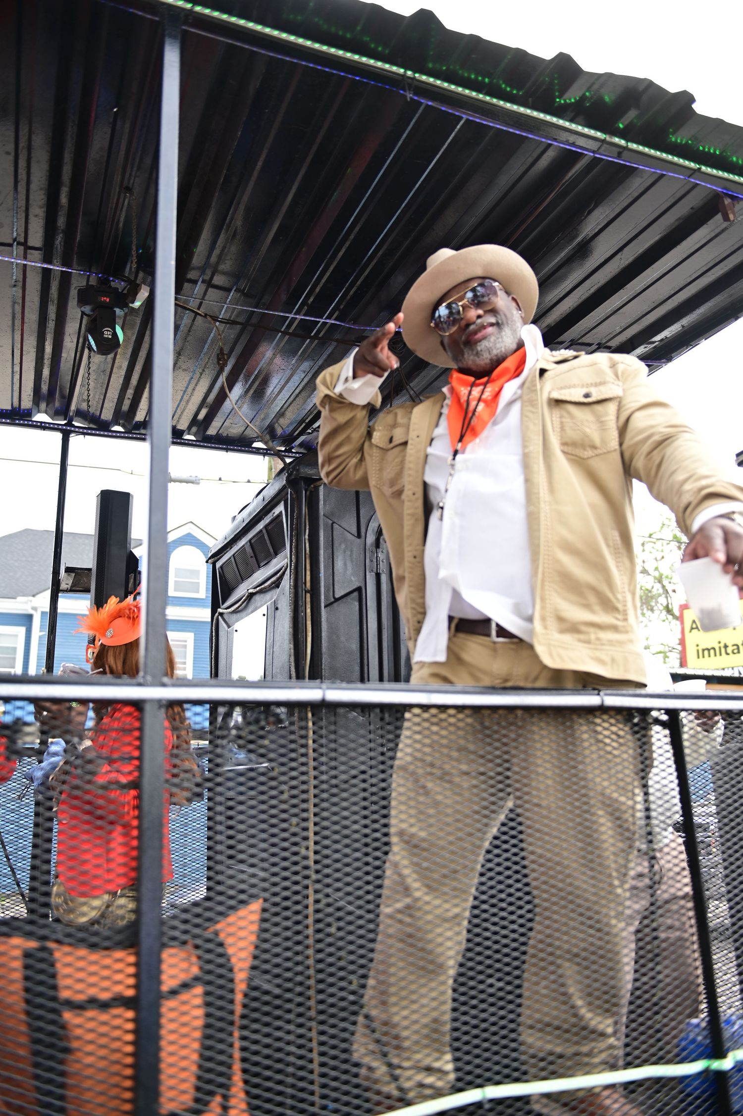 Performer in tan jacket and hat speaks into microphone on elevated platform.