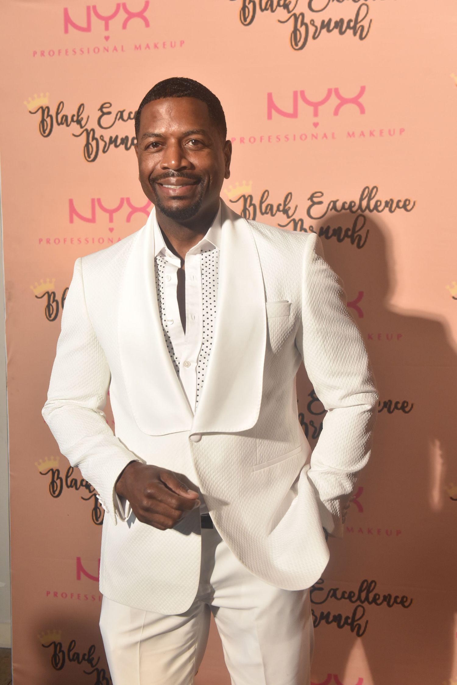 Man in white suit smiling at NYX Professional Makeup Black Excellence Brunch red carpet event.
