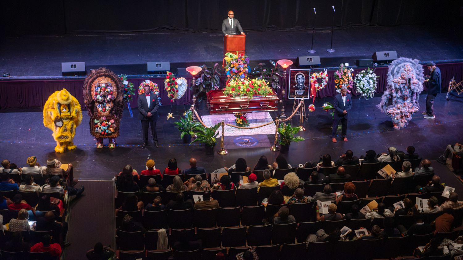A funeral service held in an auditorium with floral arrangements, a casket, and mourners seated in attendance.