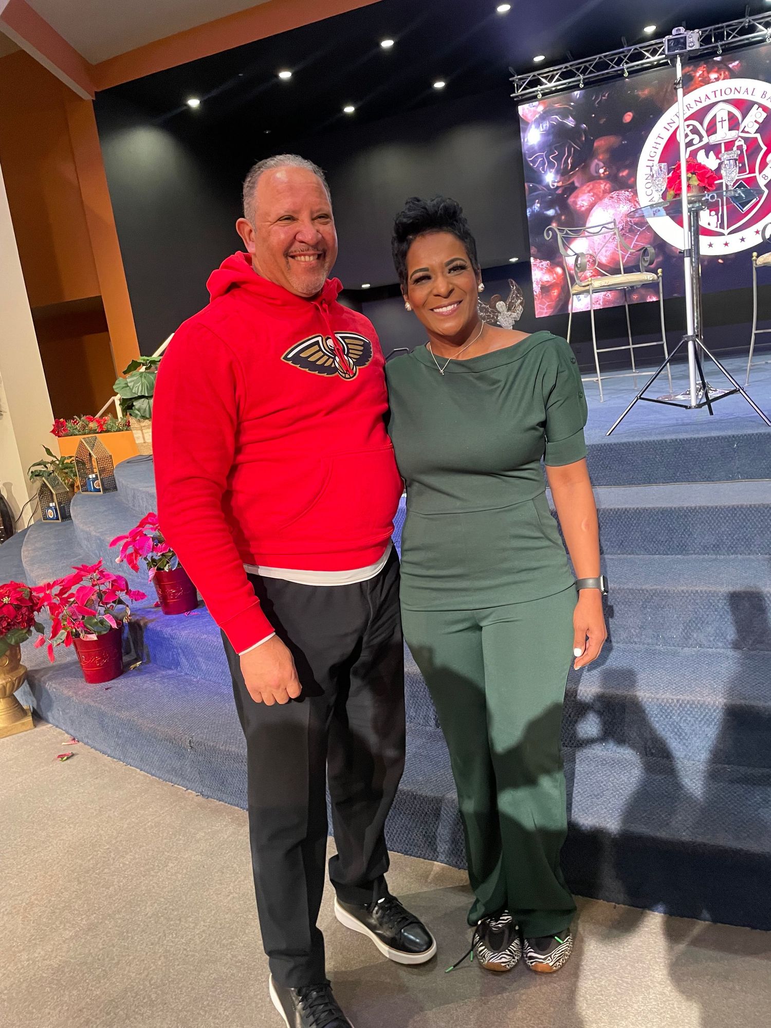 Two people in red and green outfits pose together in front of a stage area.