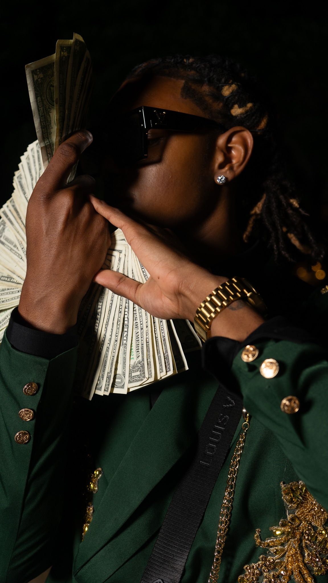 Young man in green Louis Vuitton jacket holding cash fan, wearing gold watch and sunglasses with dreadlocks.