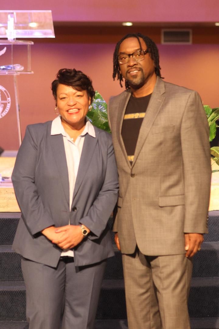 Two people in formal business attire stand together on a carpeted stage.