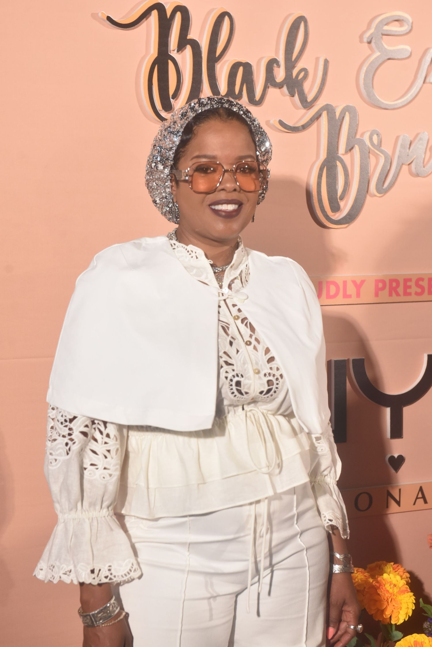 Woman in white lace outfit and glitter headscarf poses at Black Excellence Brunch event backdrop.