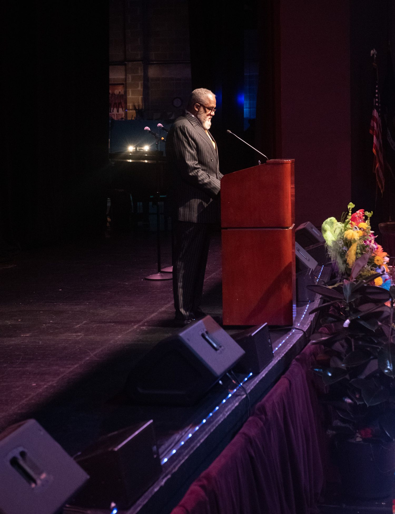 Man speaking at a red podium on a dark stage with floral arrangements and speakers in the foreground.