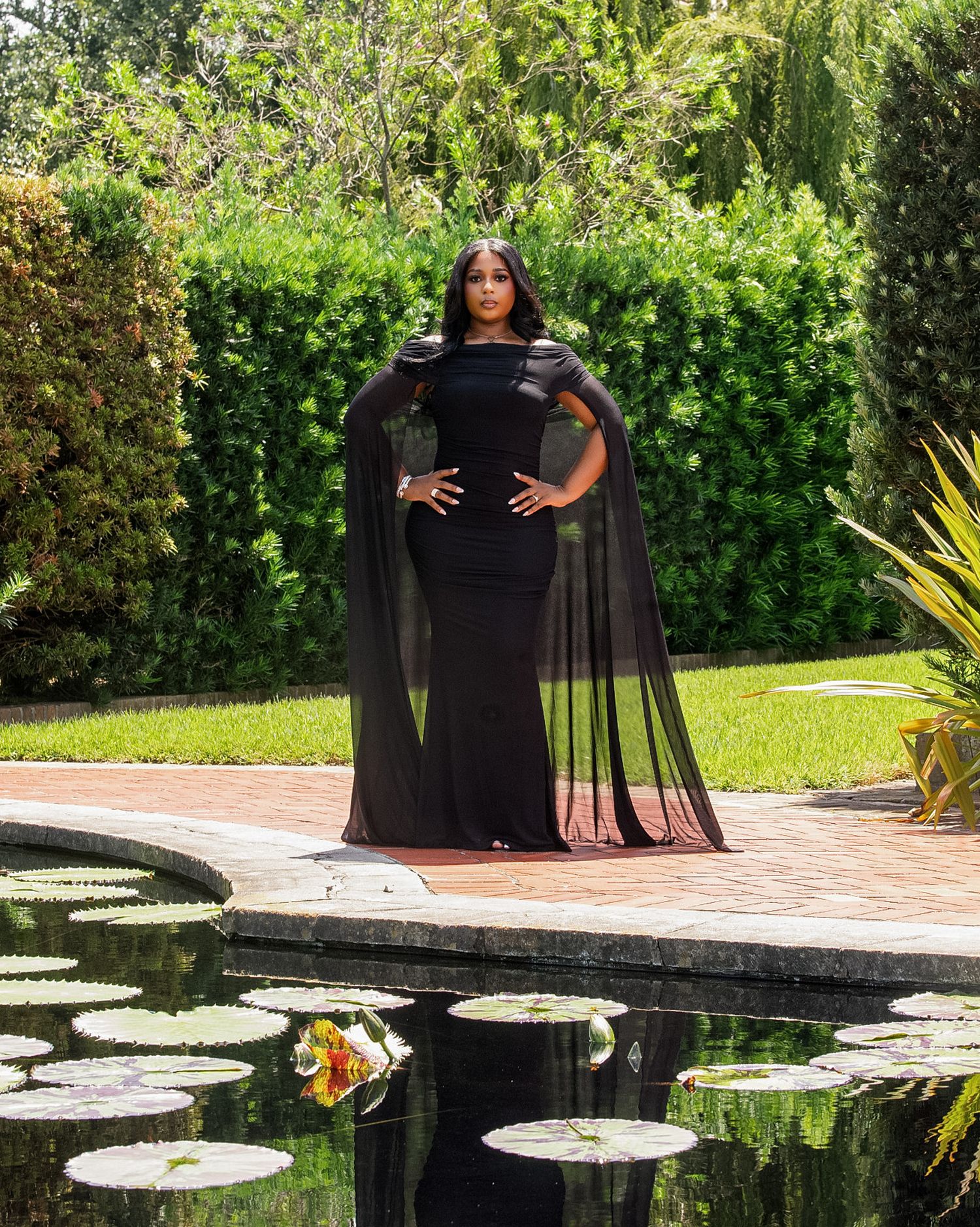 Woman in elegant black cape gown poses gracefully by a lily pond in a lush garden setting.