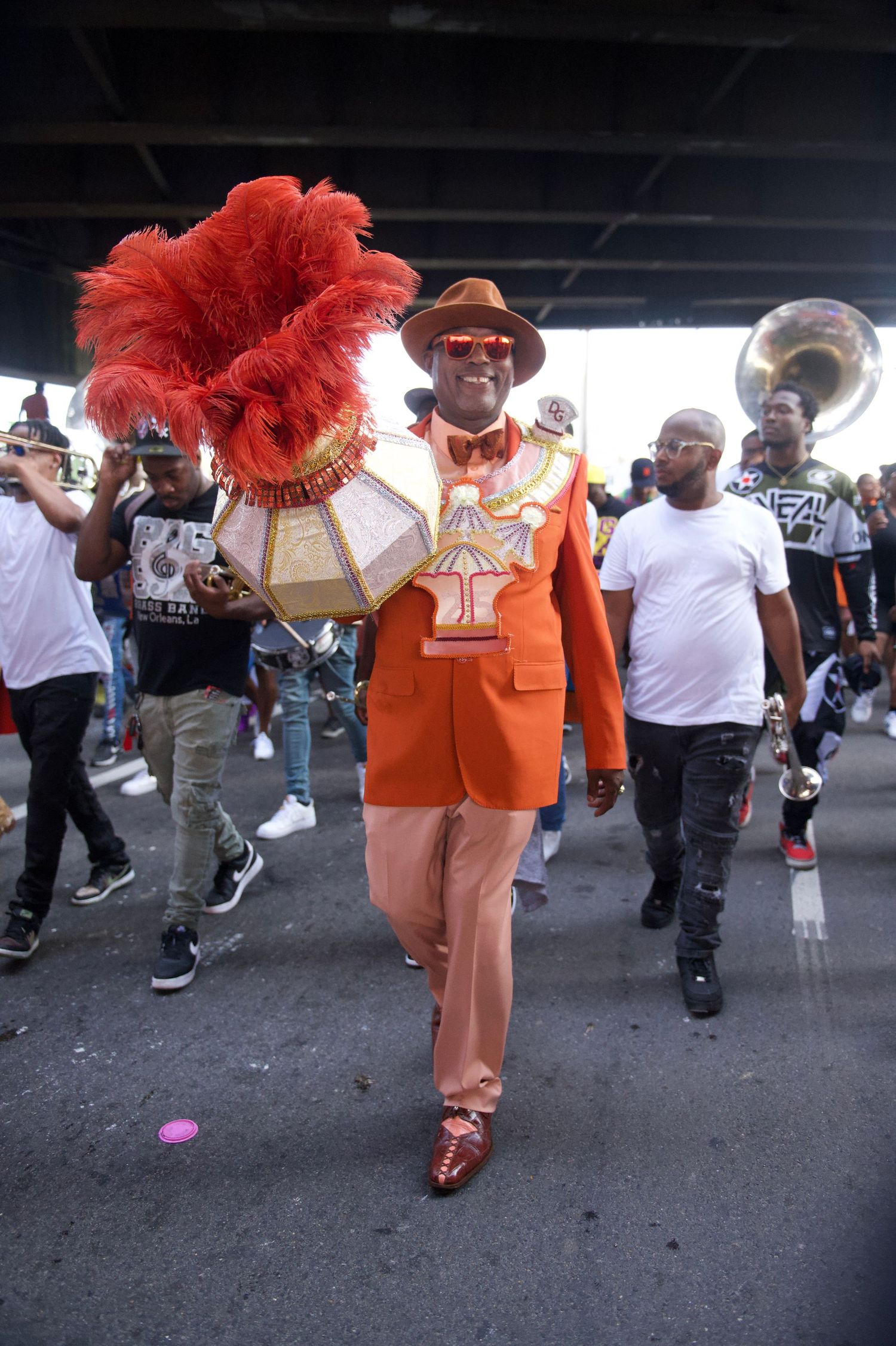 Street performer in orange suit with dramatic red feather headdress leading parade participants.