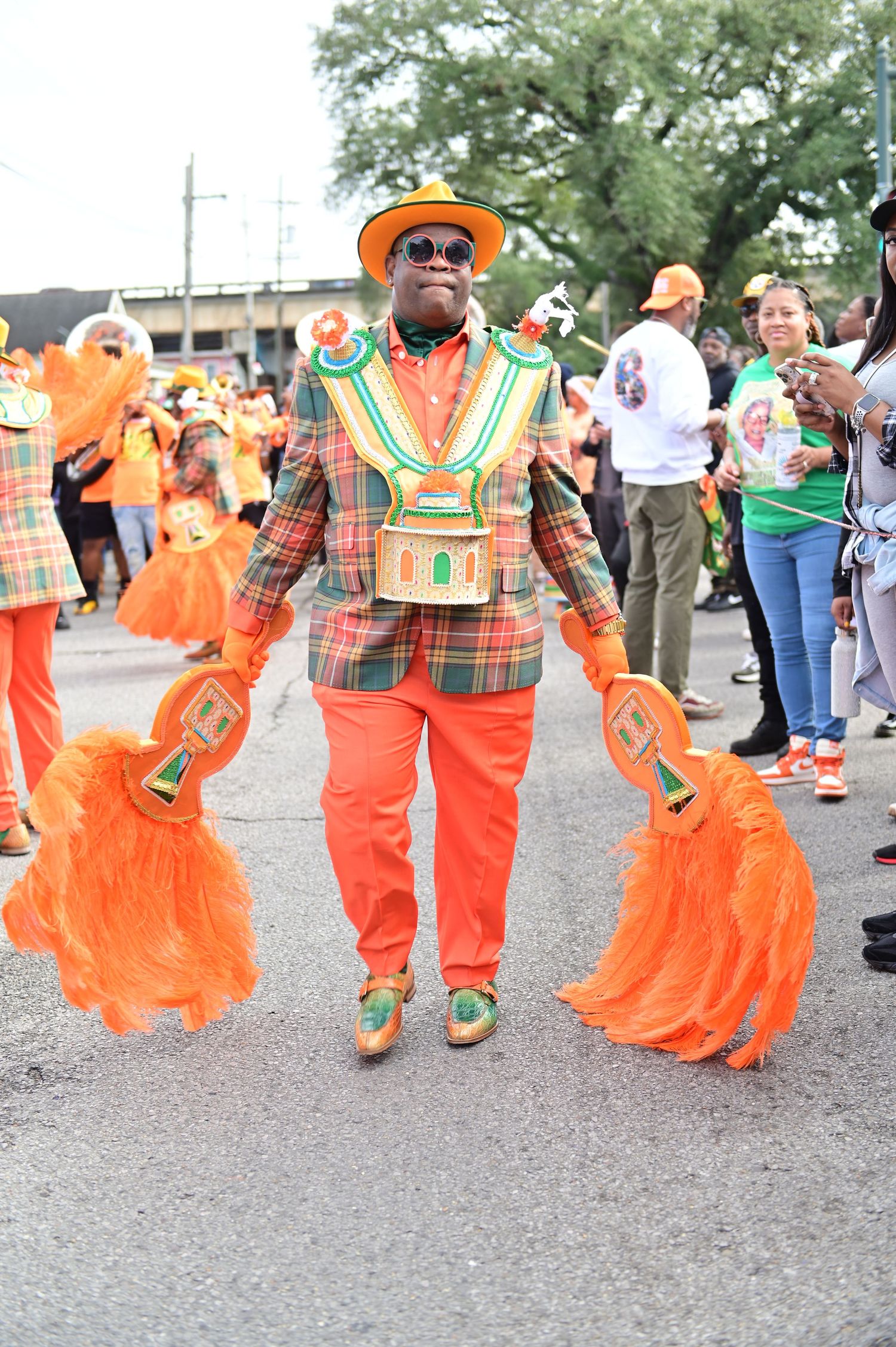 Performer holds orange feather props while wearing plaid suit and matching hat during parade.