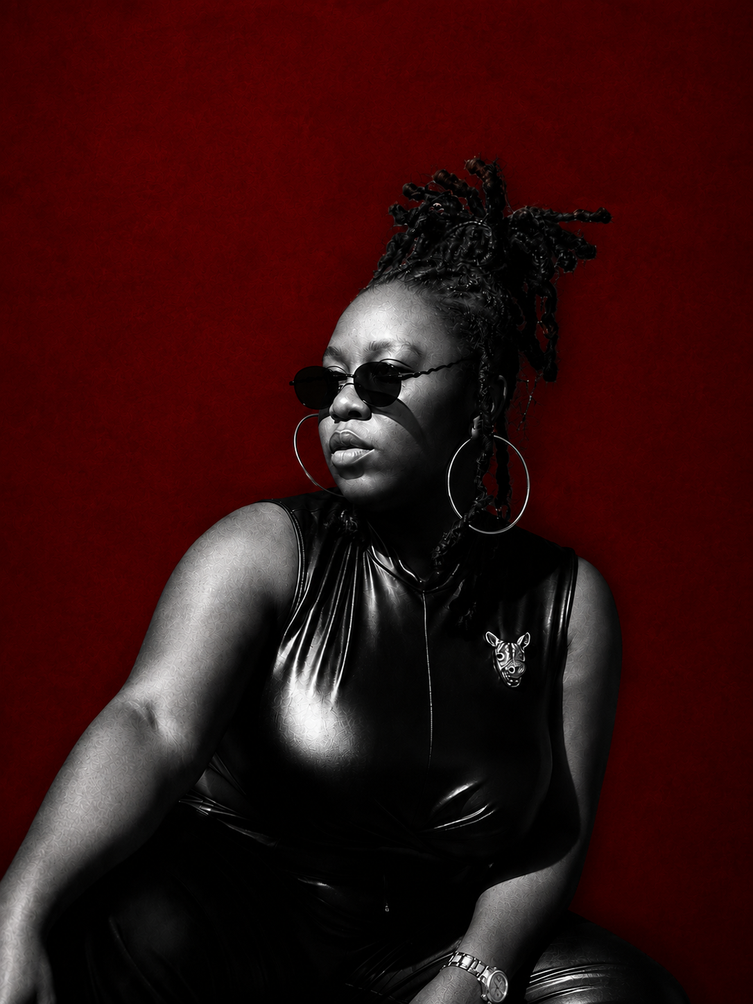 Black and white portrait of a woman in leather top with hoop earrings and sunglasses against red background.