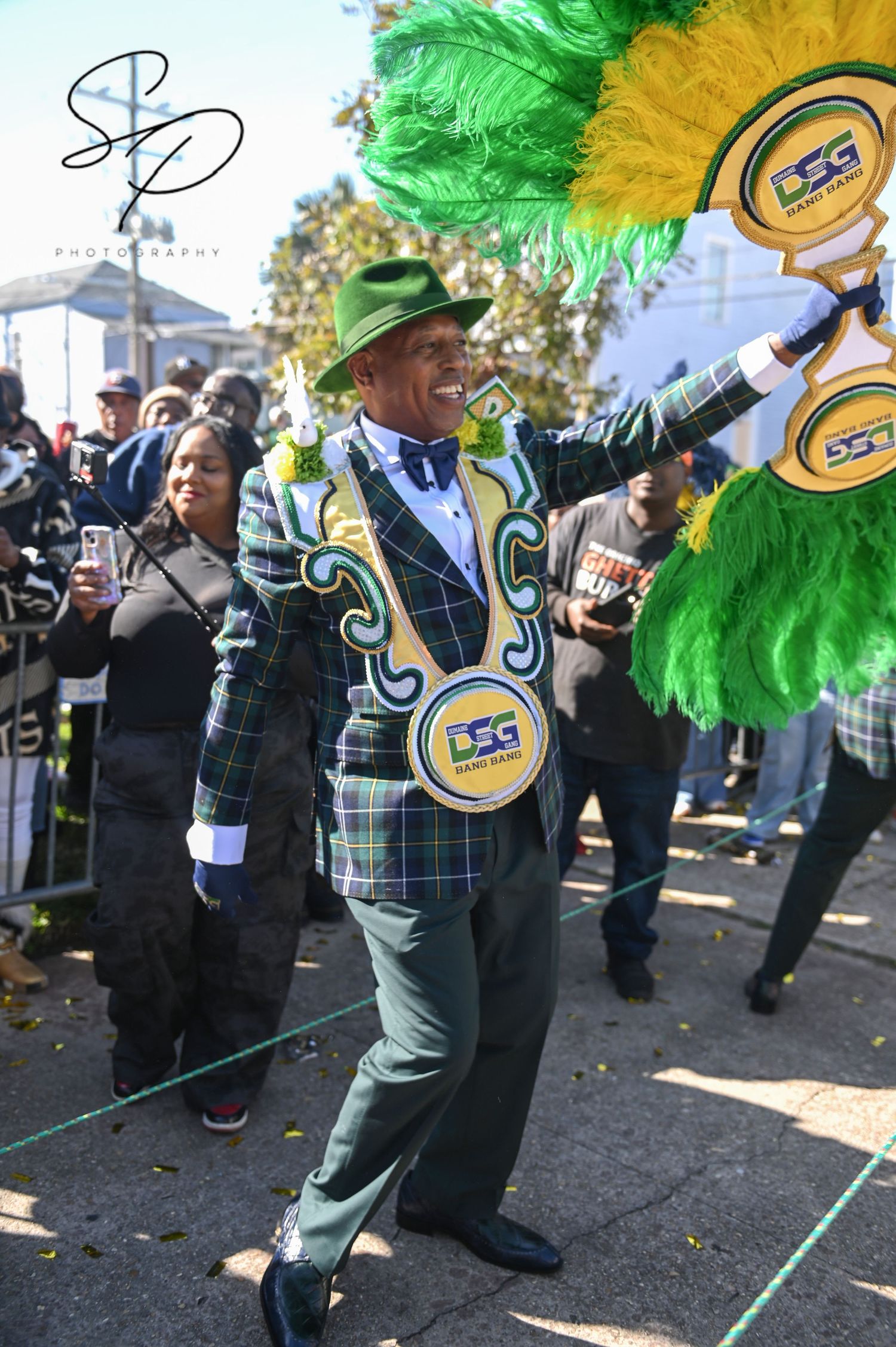 Performer in plaid suit with green and gold feathers dancing during street celebration.