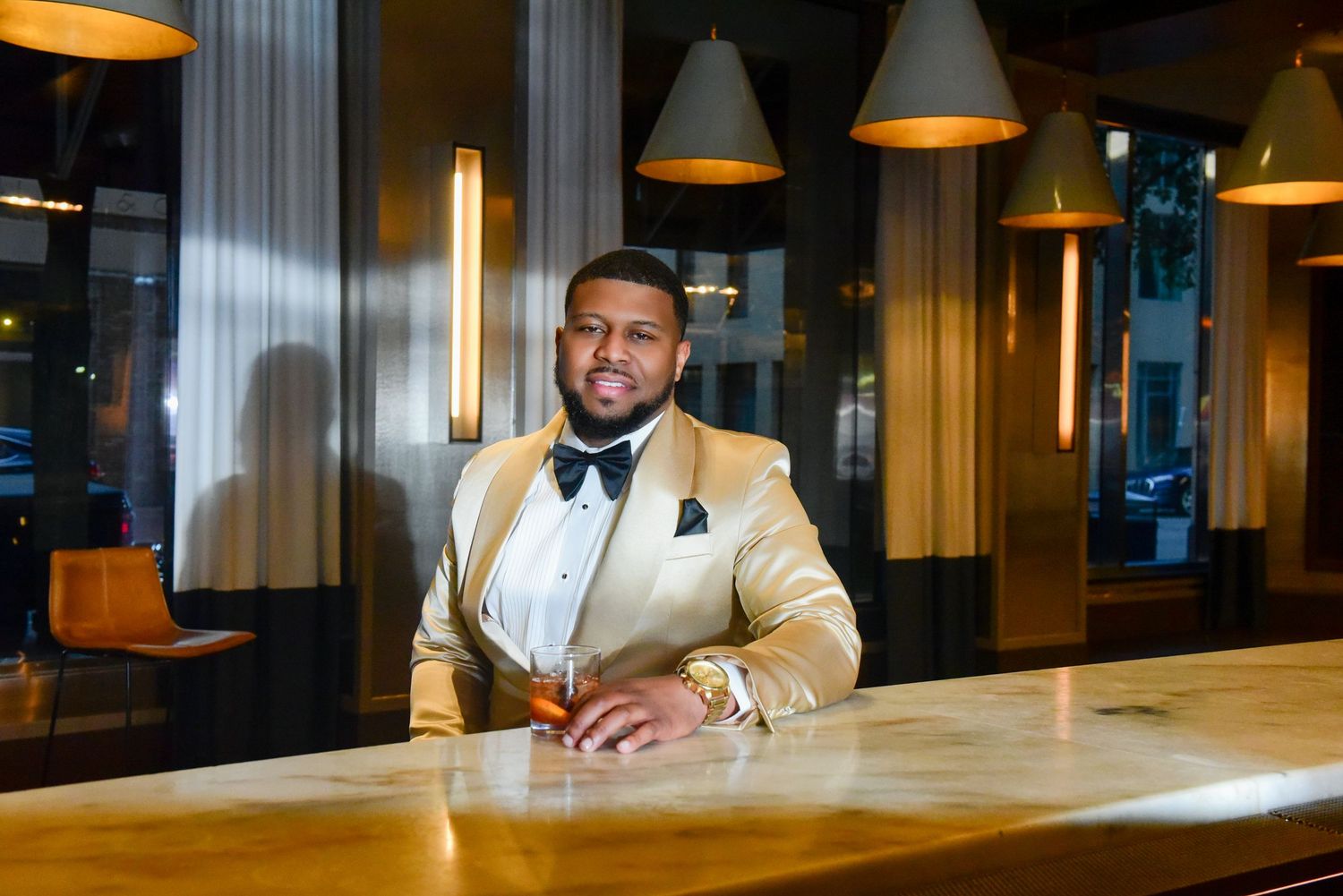 Stylish man in gold tuxedo with bow tie sitting at a bar holding a drink under warm pendant lights.