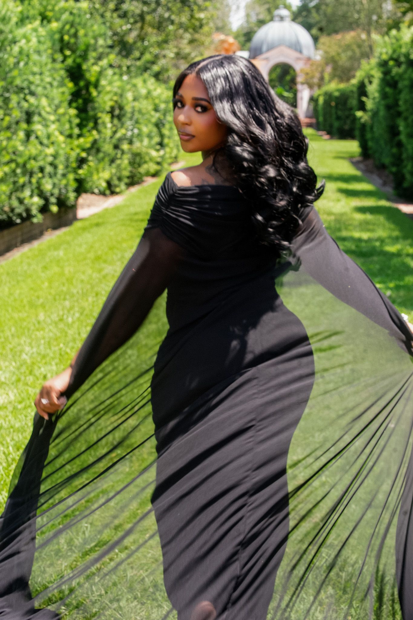 Woman in elegant black cape gown posing in lush garden.