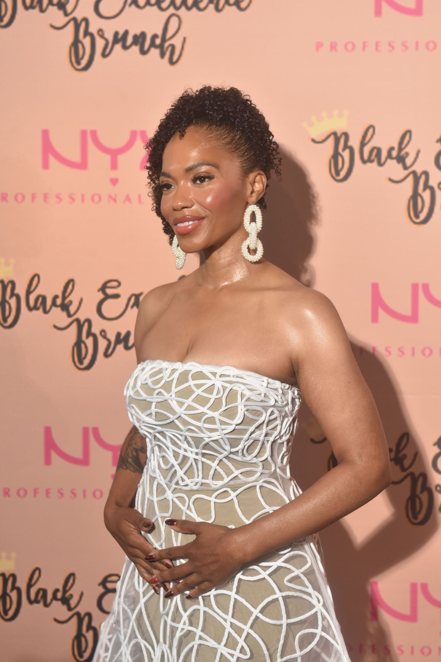 Pregnant woman in white strapless gown poses at NYX Professional Makeup Black Excellence Brunch event.