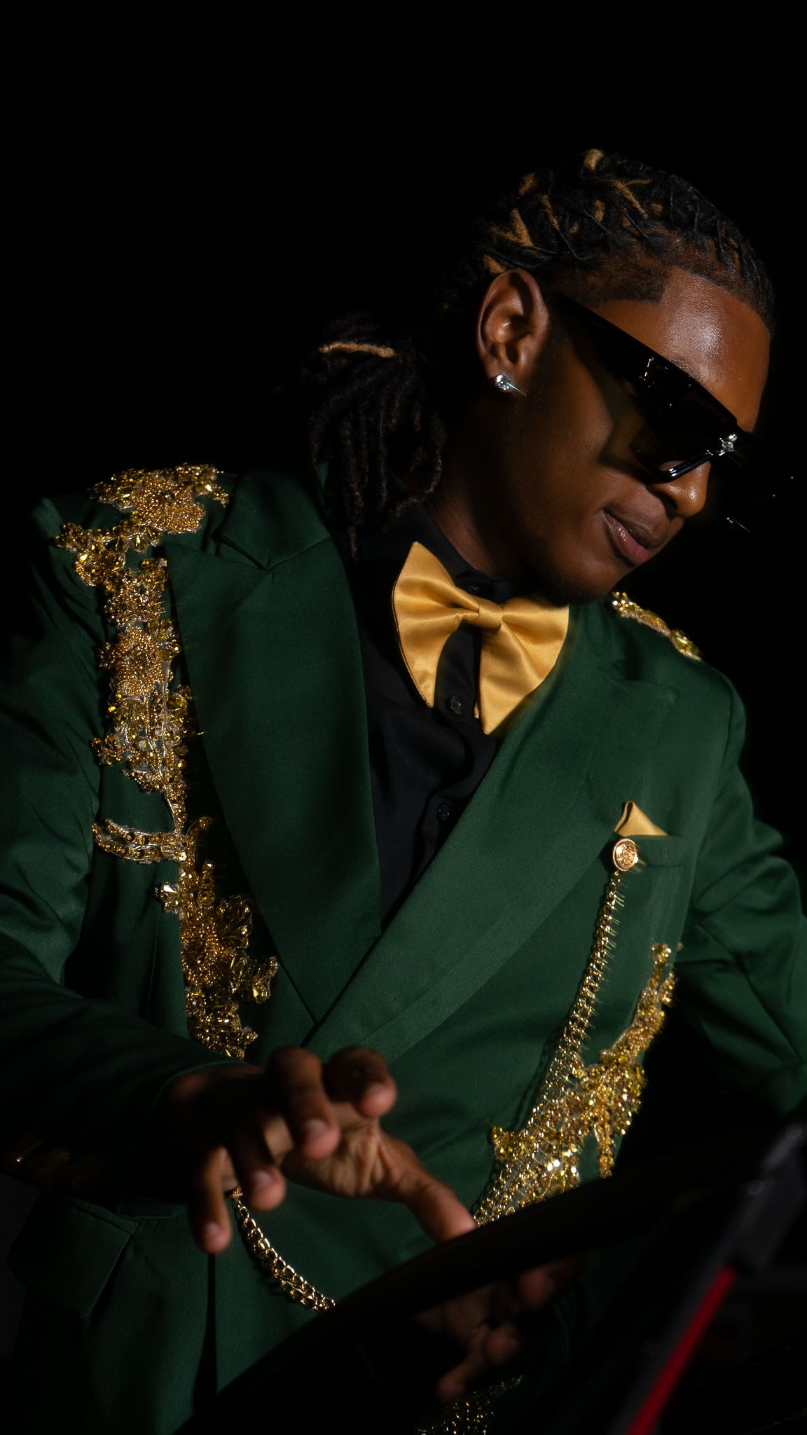 Musician in green gold-embroidered jacket, gold bow tie, sunglasses, playing steel pan drums against black background.