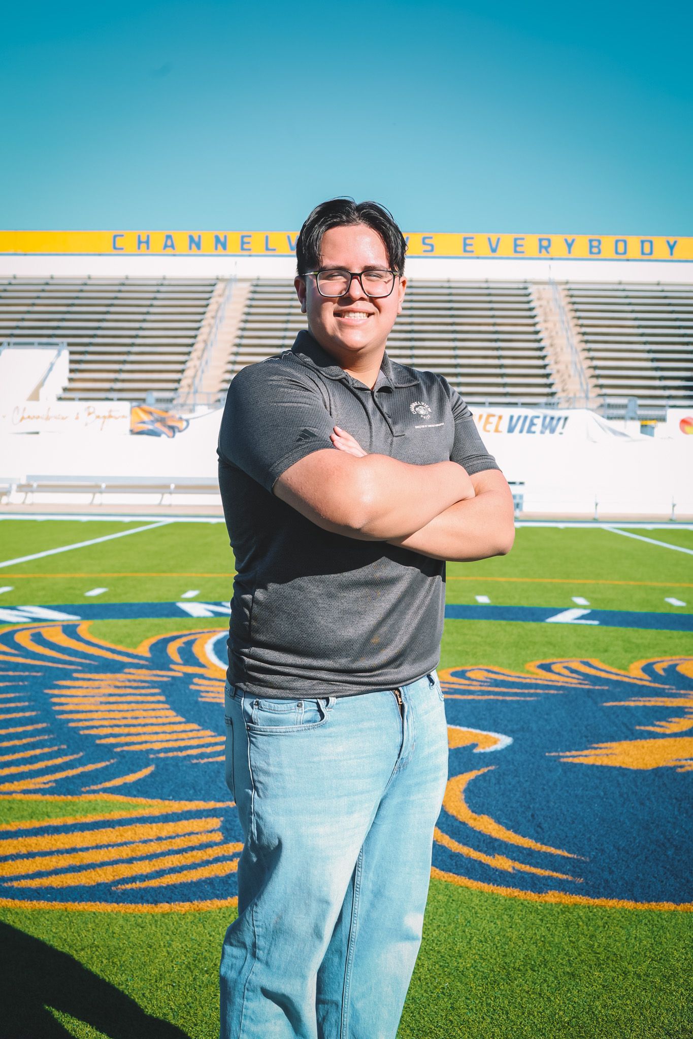 Athletic staff member stands confidently on football field with stadium bleachers and blue sky in background.