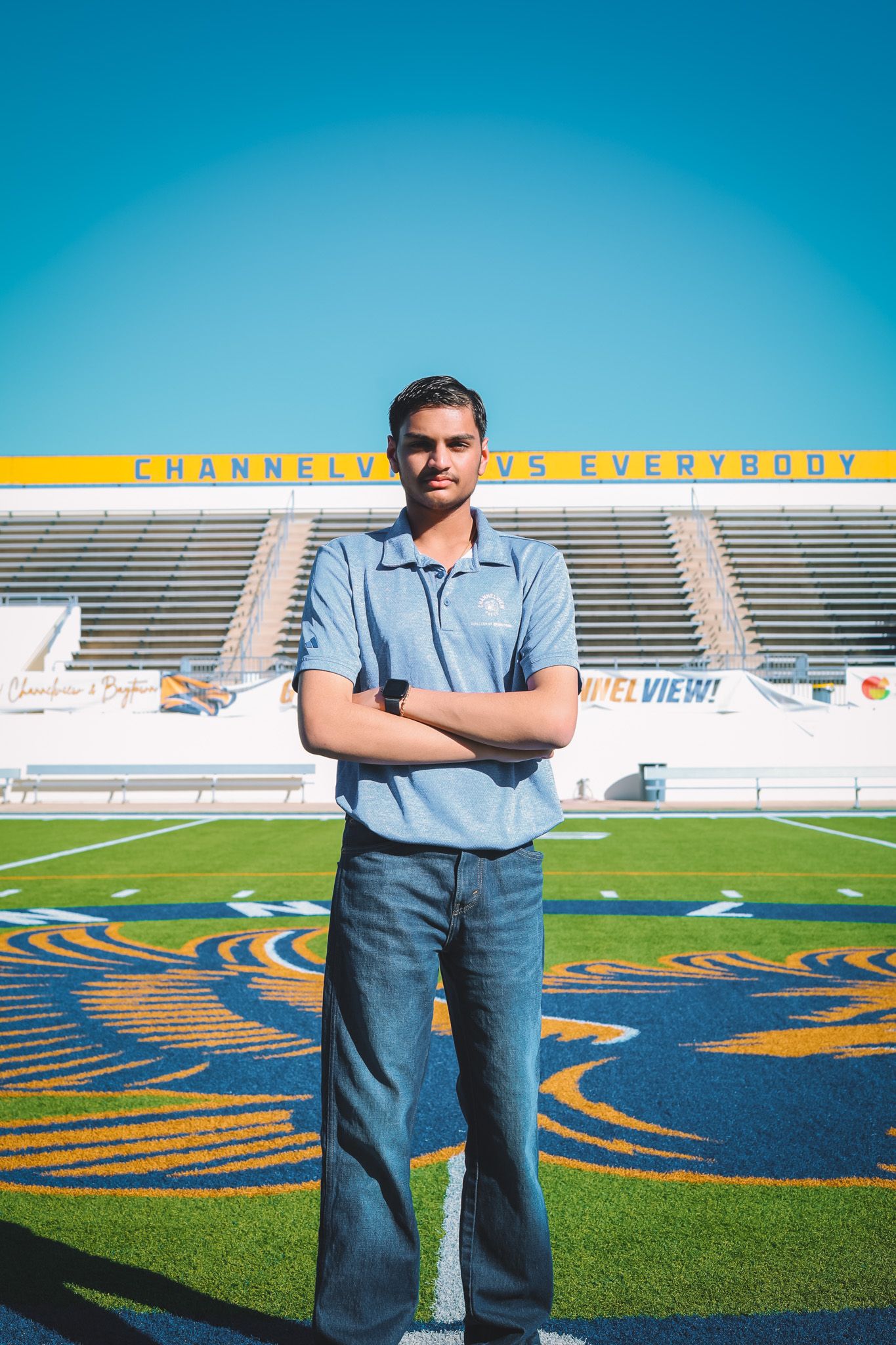 Person standing on football field with stadium bleachers and blue sky in background.