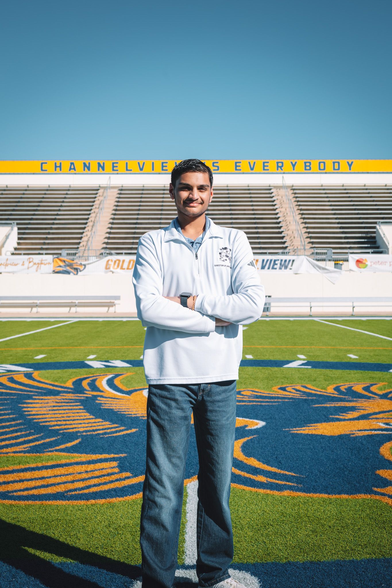 Young man in white pullover stands arms crossed on football field at Channelview stadium on a sunny day.